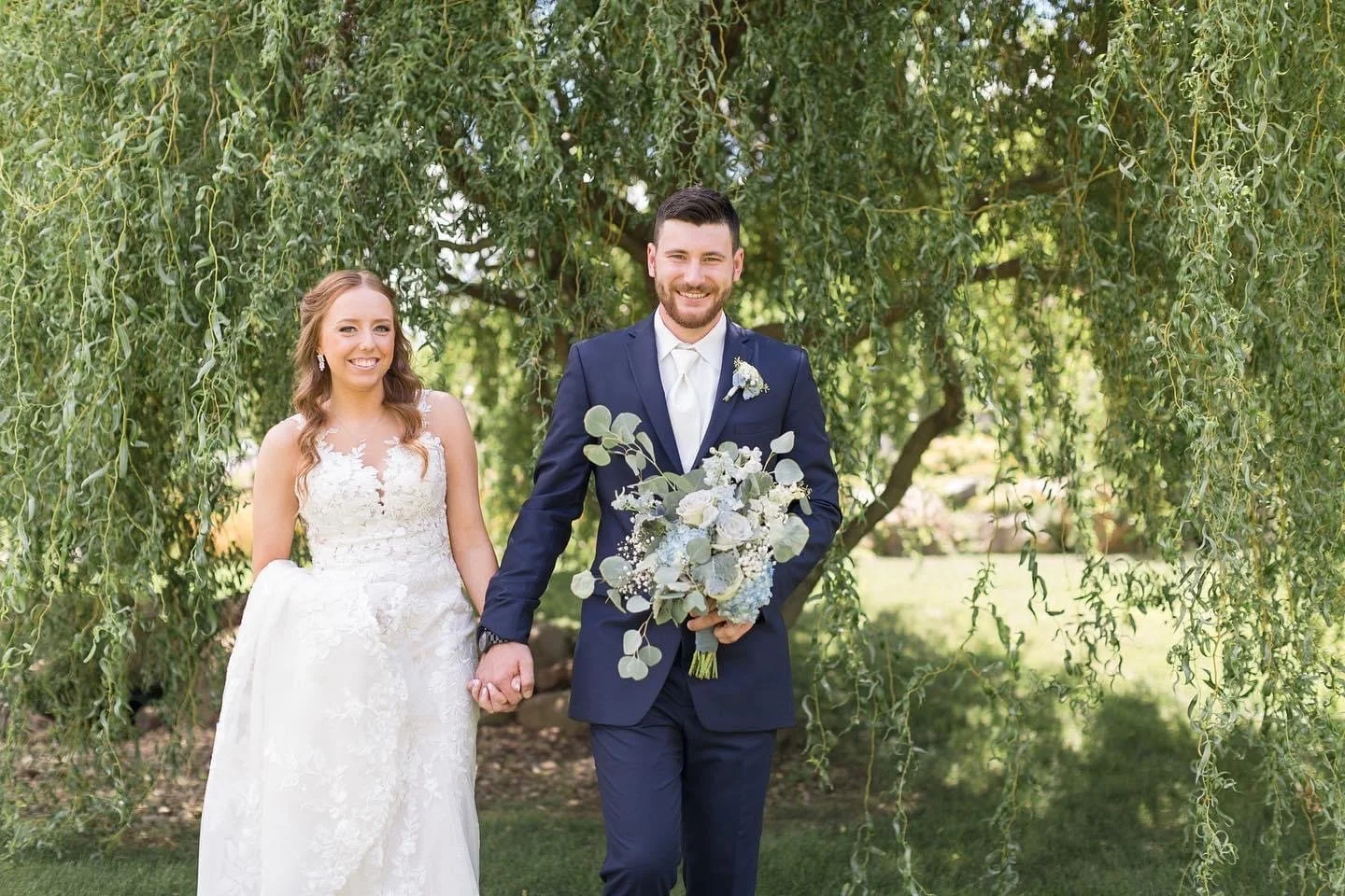 Bride and groom holding hands outdoors beneath leafy tree, smiling, wedding attire, bride in white lace gown, groom in navy suit, holding bouquet of white roses and greenery.