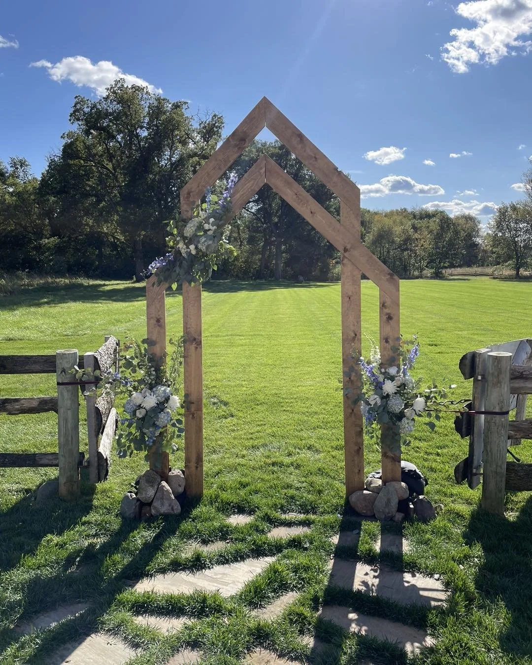 Wooden arch decorated with flowers and rocks in a grassy outdoor setting with a blue sky and trees in the background.