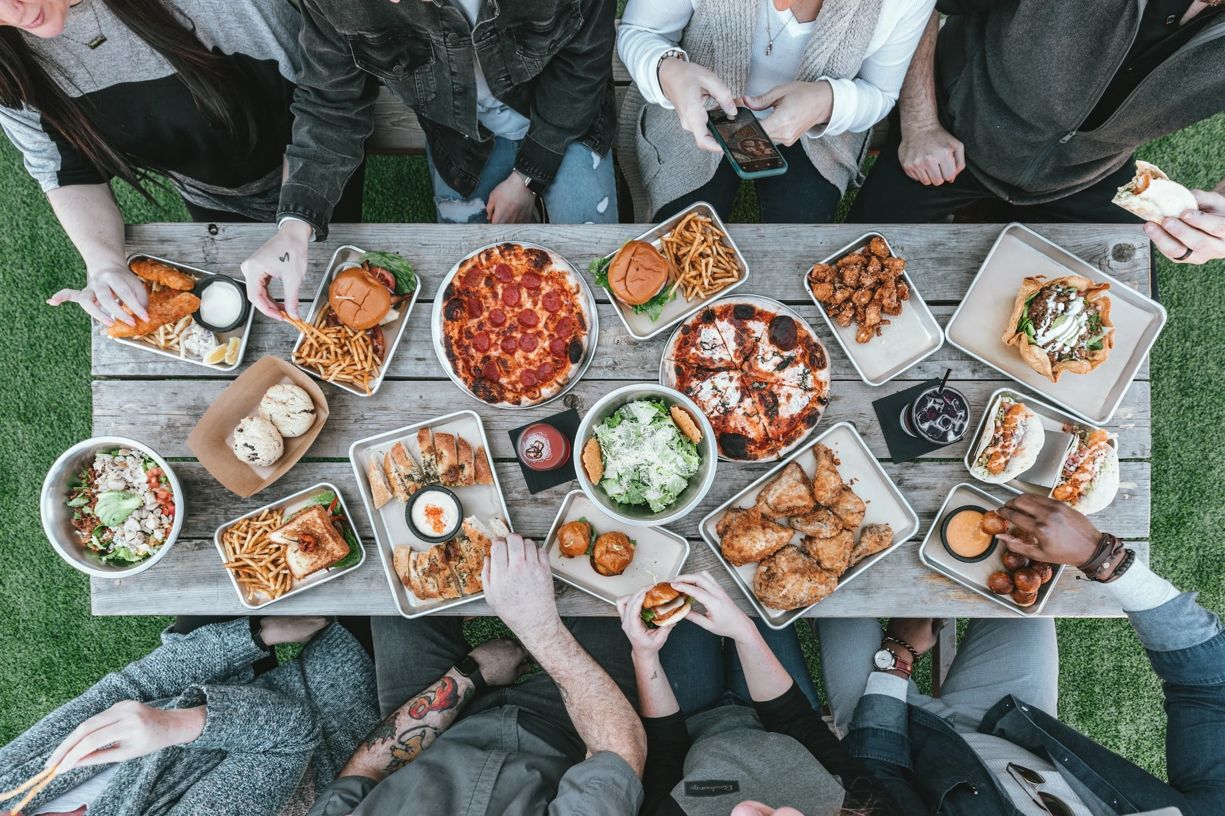 A top-down view of a group of people at a wooden outdoor table filled with various plates of food including pizza, sandwiches, fried chicken, salad, French fries, and drinks.