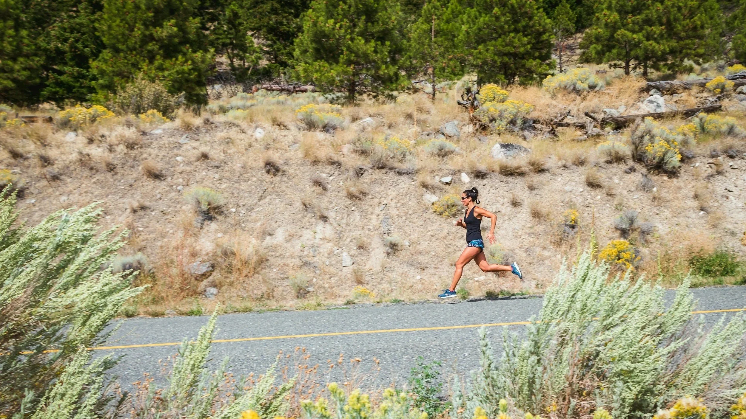 A woman running along a paved road in a dry, hilly landscape with green bushes and trees.