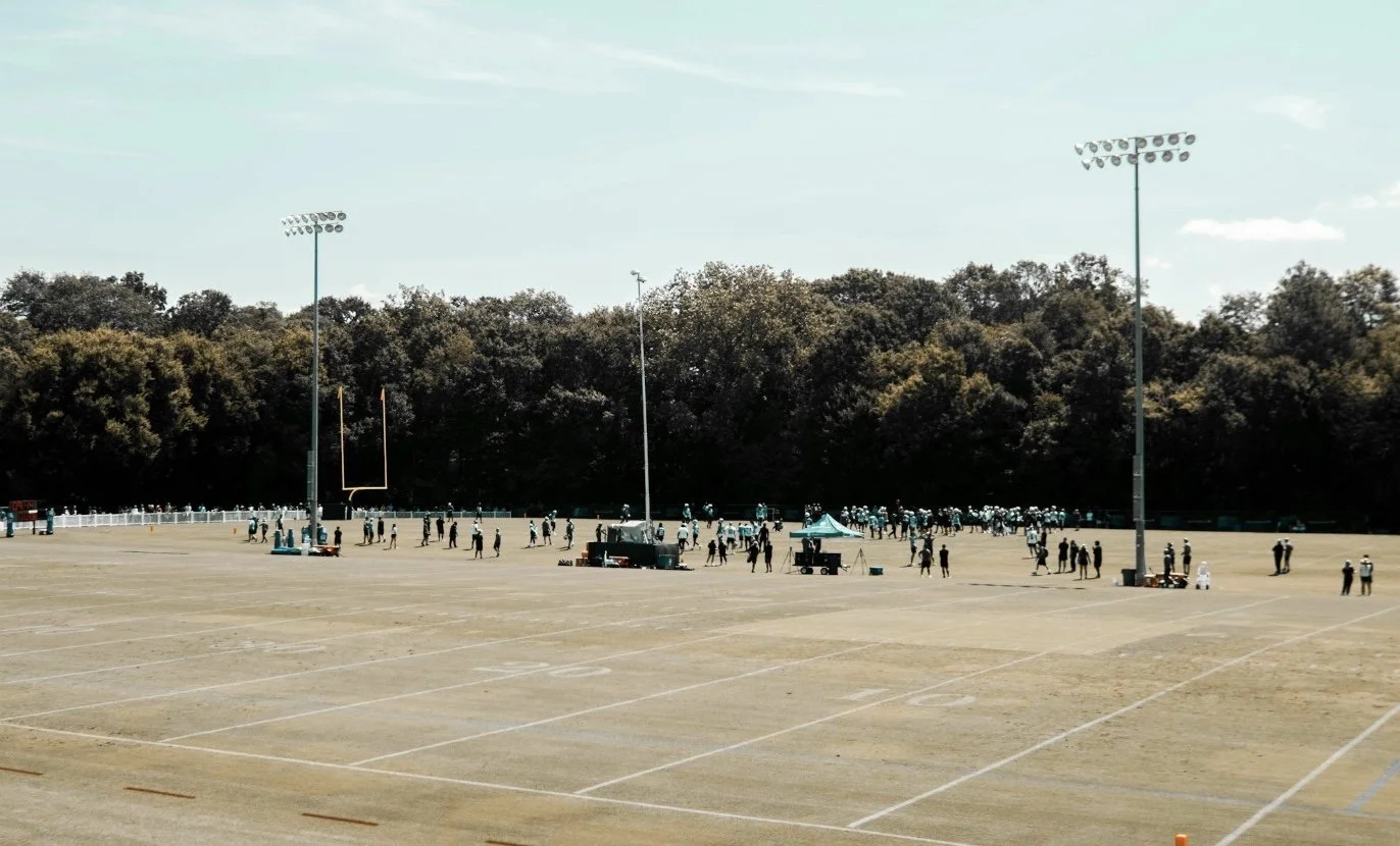 Wide view of a football field with people gathered, tents, and tall light poles, with trees in the background on a clear day.