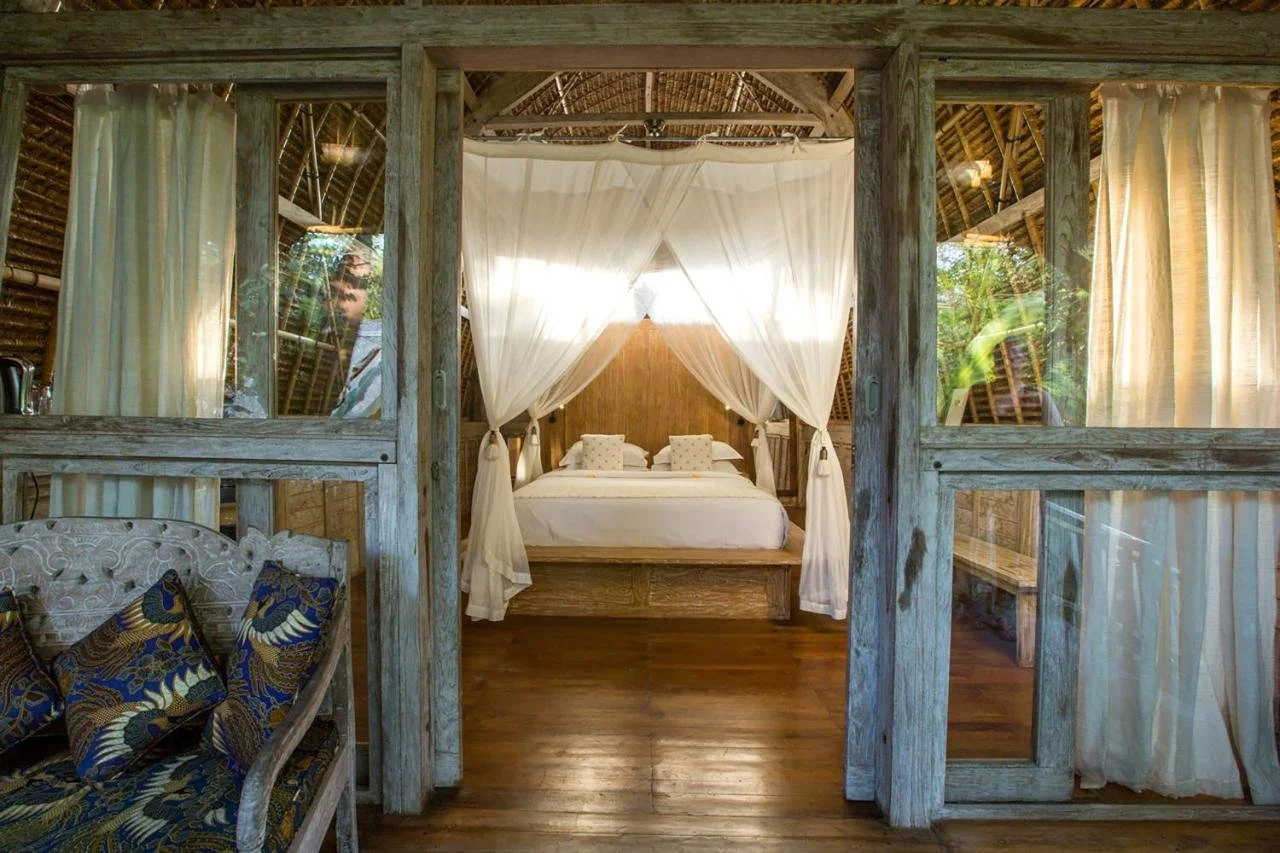Wooden interior of a tropical bungalow bedroom with a four-poster bed draped with white curtains, framed by glass-paneled wooden walls and curtains, with a sitting area and lush greenery visible outside.