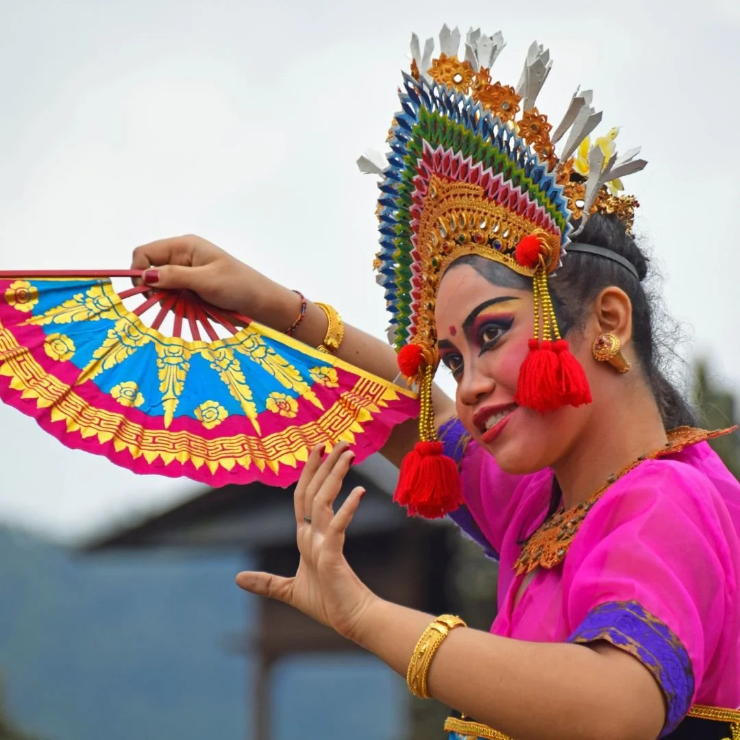 A woman dressed in traditional Indonesian dancer attire, wearing a colorful headdress and pink costume, poses with a decorative pink and yellow fan.