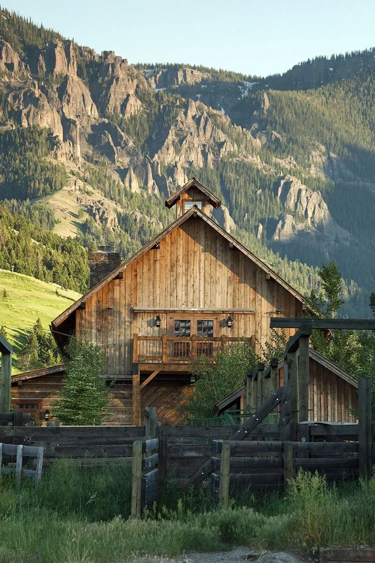 Wooden house with a porch and railing, surrounded by trees and colorful plants, with mountains in the background under a clear blue sky.