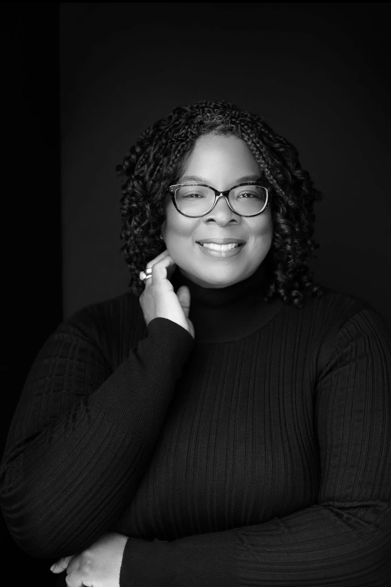 A oval shaped black and white photo of Amber Henderson, a Black woman with shoulder-lenth black hair. She is wearing black glasses and a black sweater. Her arm is folded across her stomach while the other hand is touching her jawline.