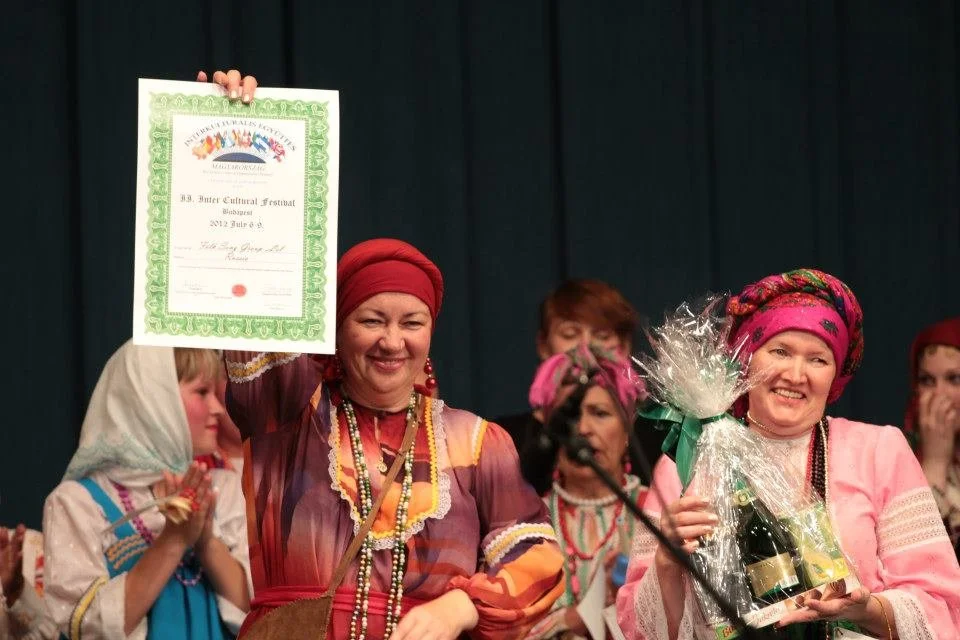 Women in traditional colorful clothing and headscarves celebrating on stage, one woman holding a framed certificate, others smiling with gifts and flowers.