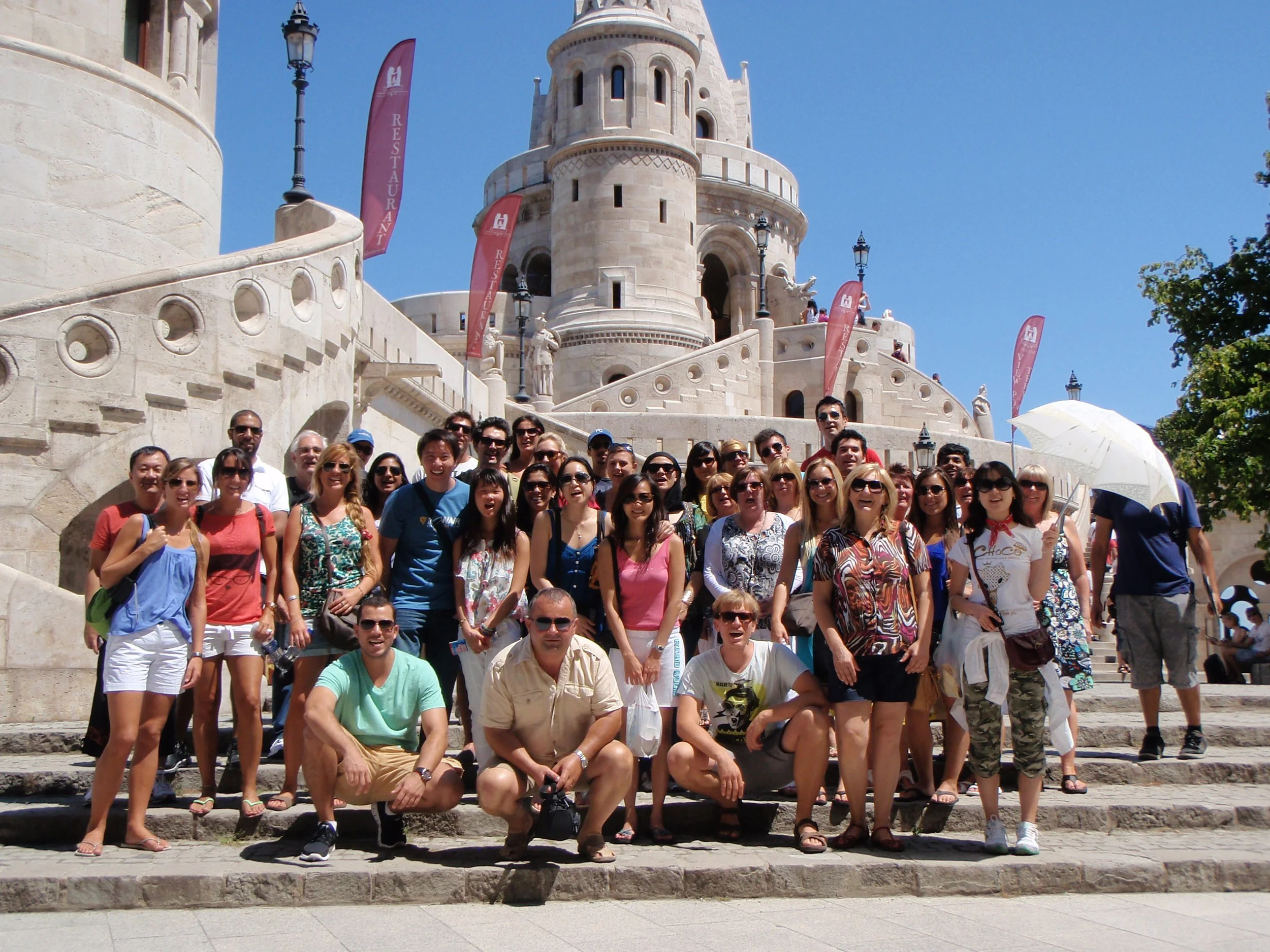 Large group of tourists posing for a photo on stone steps in front of the Fairy Tale Castle at Peles Castle in Romania. The castle features medieval architecture with pointed towers and ornate designs, under a bright blue sky with a few trees visible.
