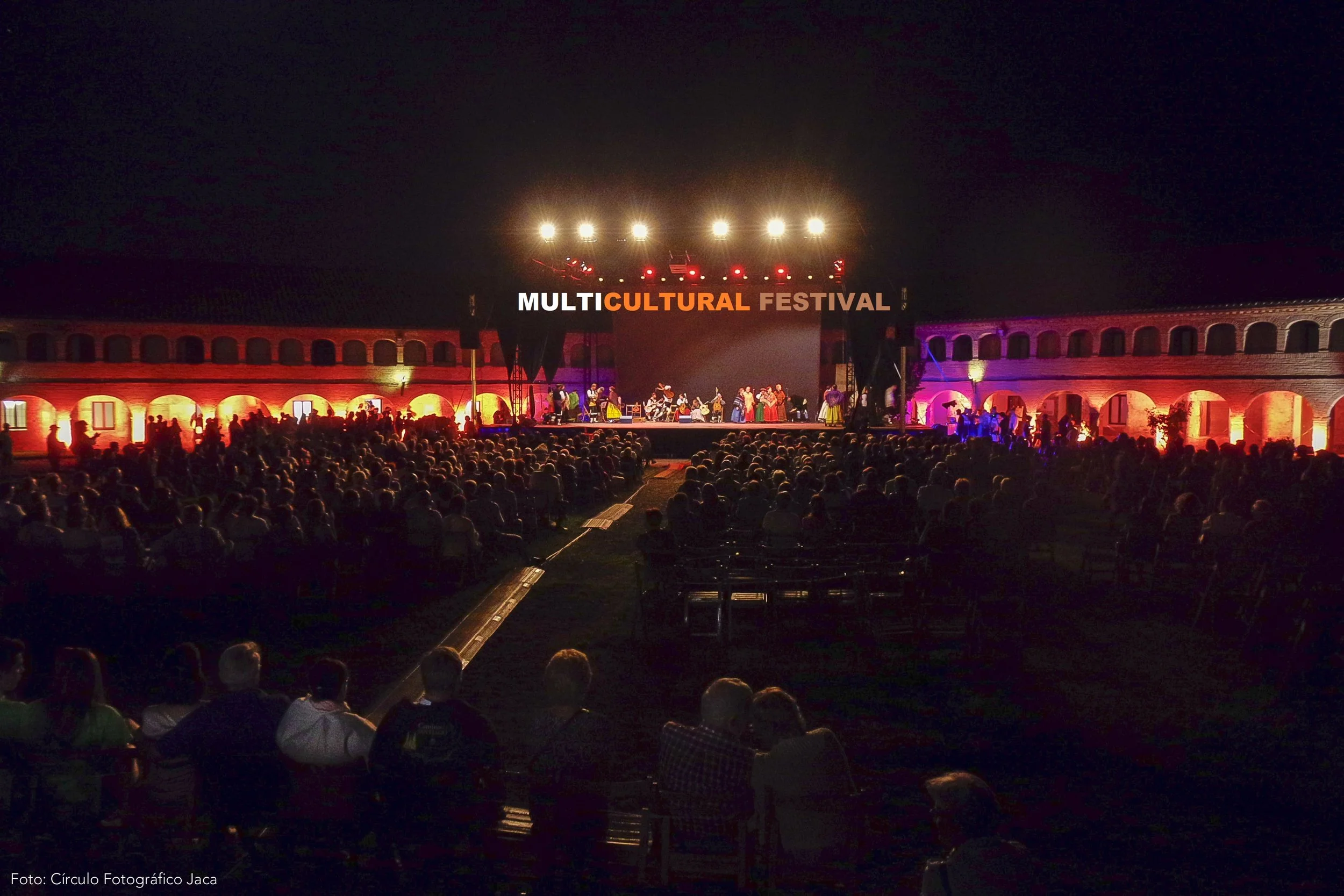 Nighttime scene of a large crowd watching a multicultural festival performance on an outdoor stage illuminated with colorful lights. The stage features a sign reading 'Multicultural Festival' and is set against an ancient-looking brick structure with arches, lit with purple and orange lights.