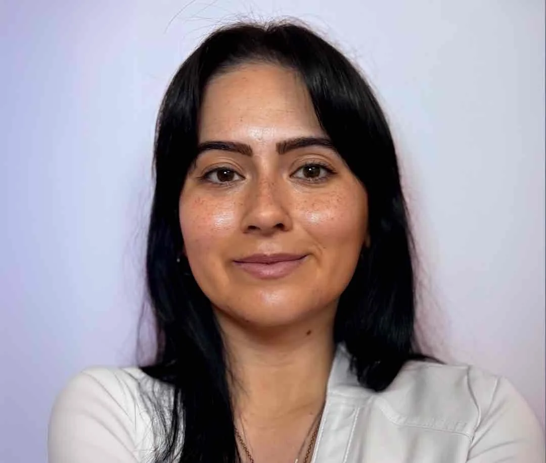A woman with medium skin, long black hair, and freckles, smiling slightly, wearing a white top, against a light-colored background.