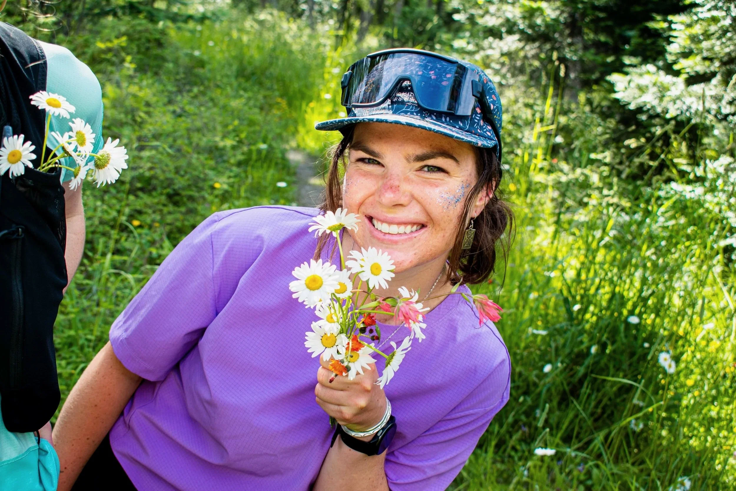 A woman with brown hair and freckles holding a bouquet of white and pink daisies, wearing a purple shirt, sunglasses on her head, and a hat, smiling in a green outdoor setting.