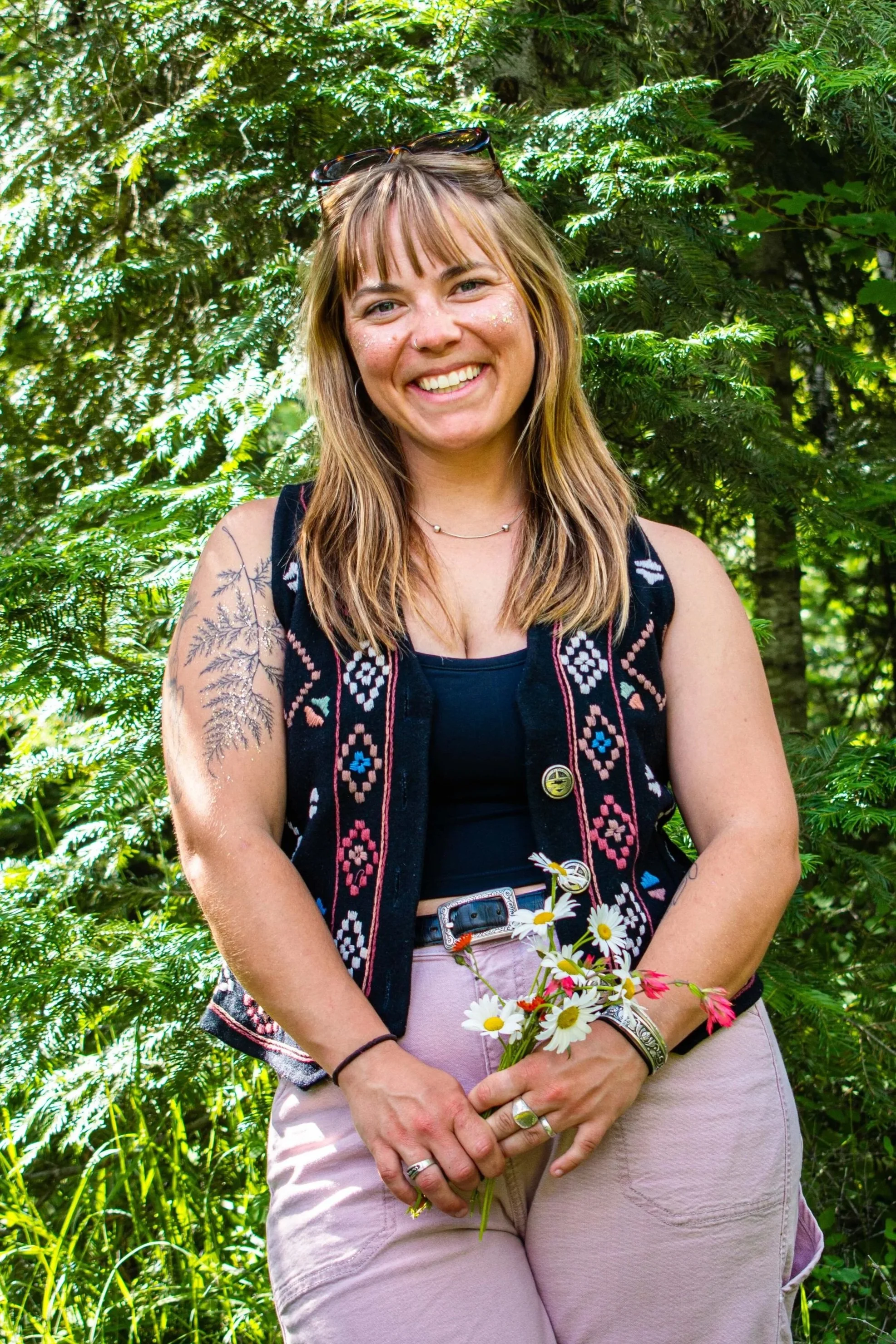 A smiling woman with shoulder-length light brown hair and bangs, wearing sunglasses on her head and a black embroidered vest, holding a bouquet of daisies, standing in front of lush green trees and plants.