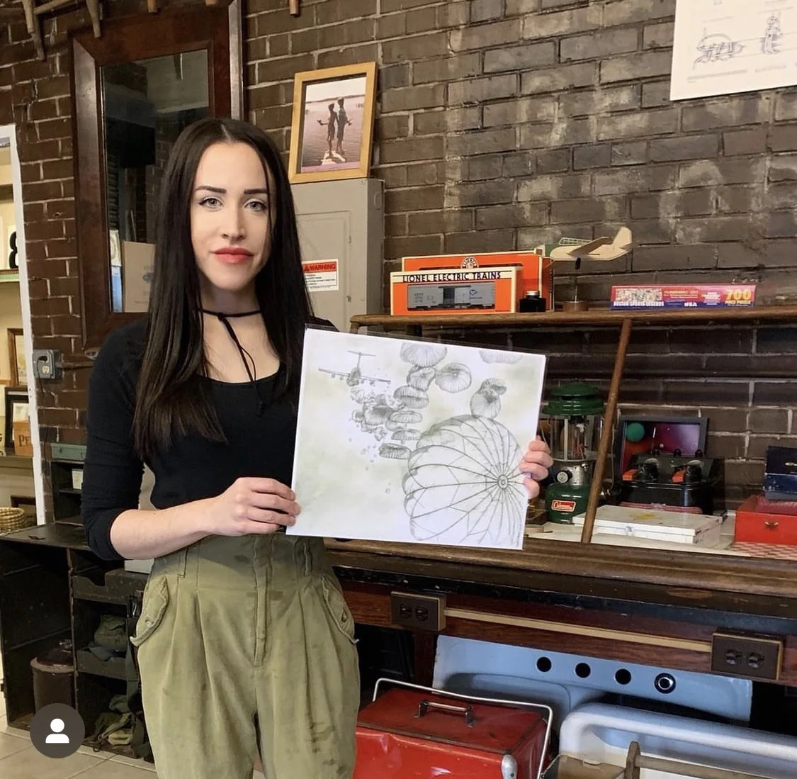 A woman with long dark hair and fair skin holding a drawing of abstract shapes and patterns. She is standing indoors near a wooden counter with various vintage items and a brick wall in the background.