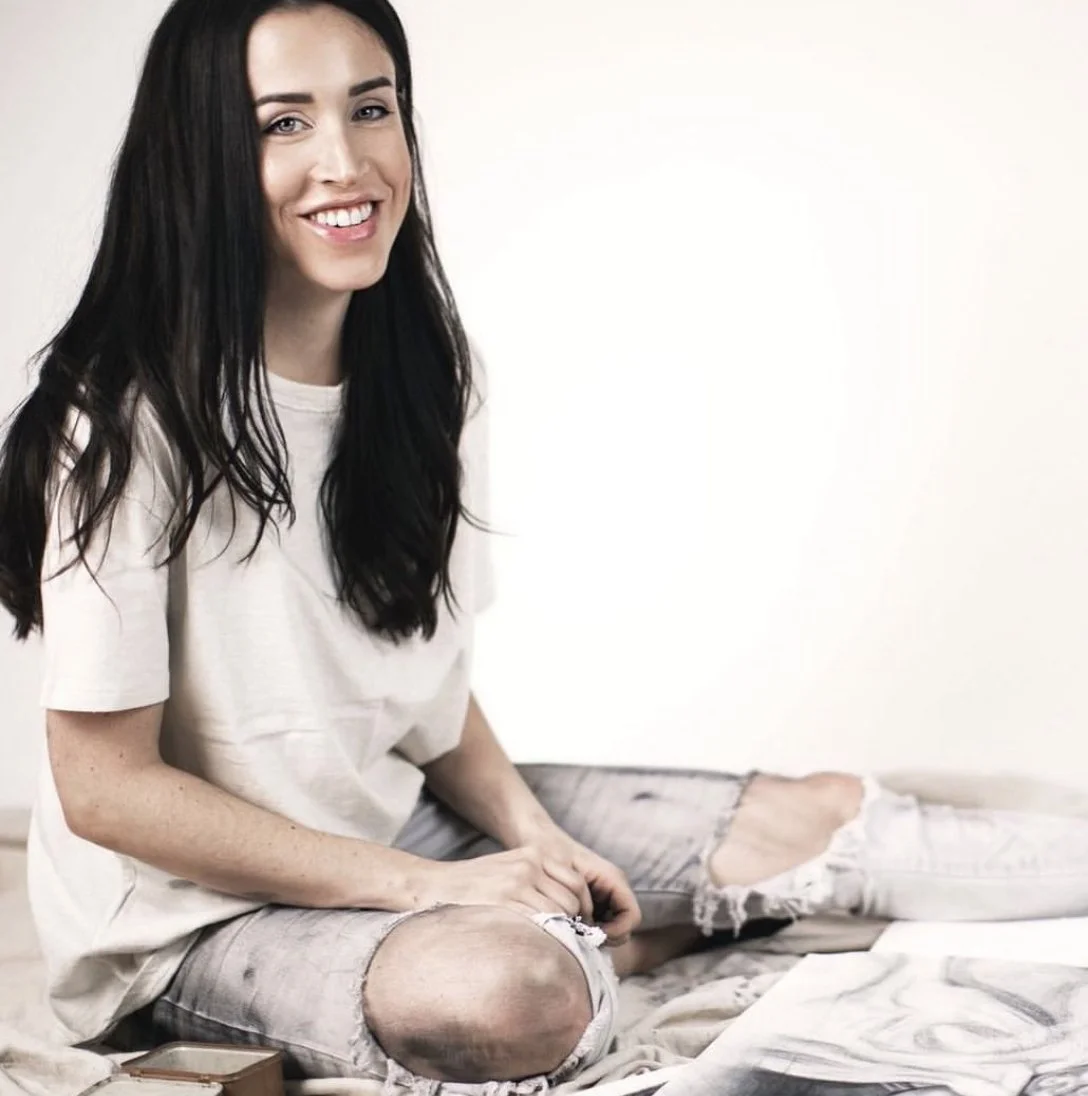A young woman with long dark hair and a bright smile, sitting on the floor with her legs crossed. She is wearing a white t-shirt and ripped jeans, in front of a plain white background.