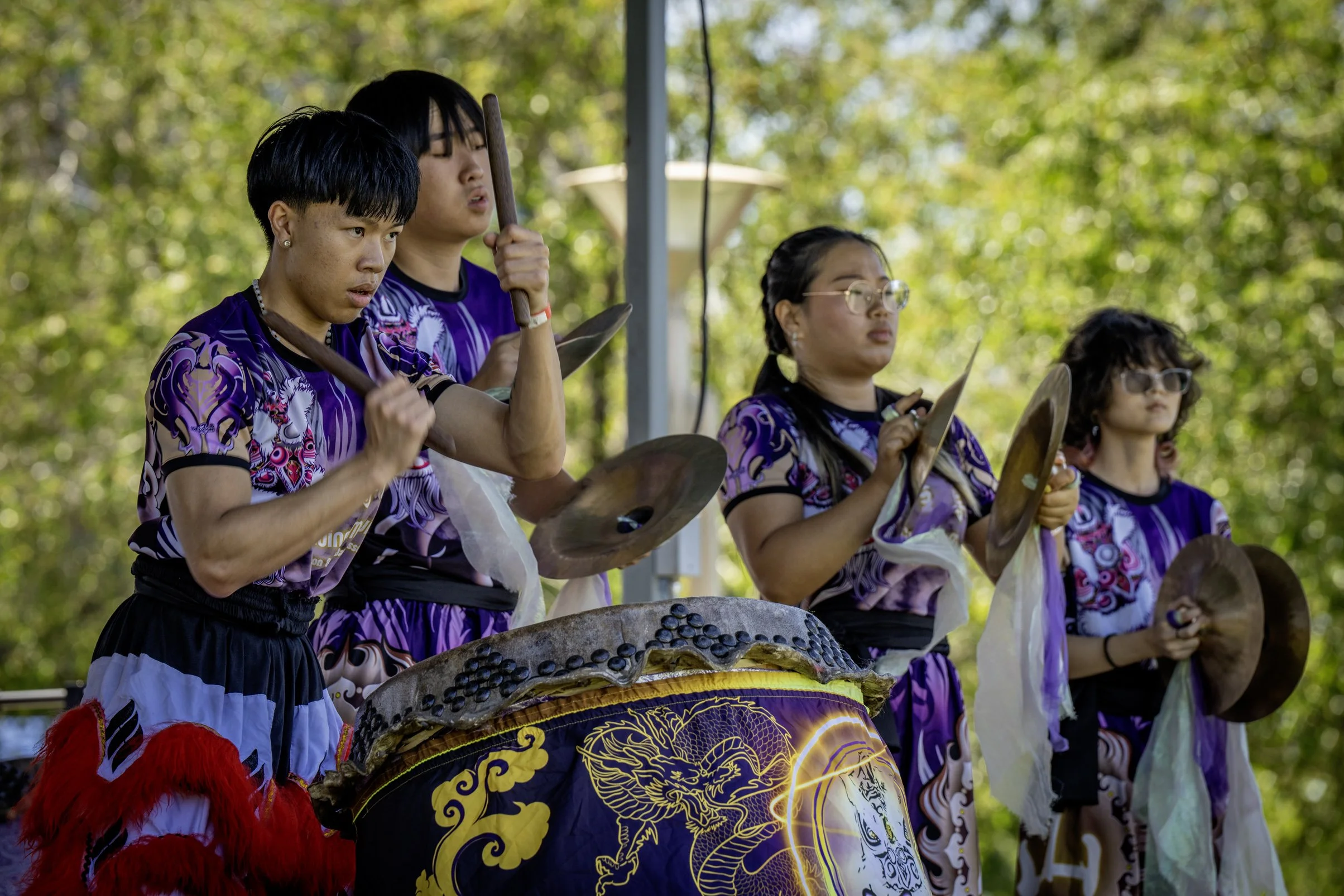 Four young people wearing purple costumes playing traditional percussion instruments outdoors with trees in the background.