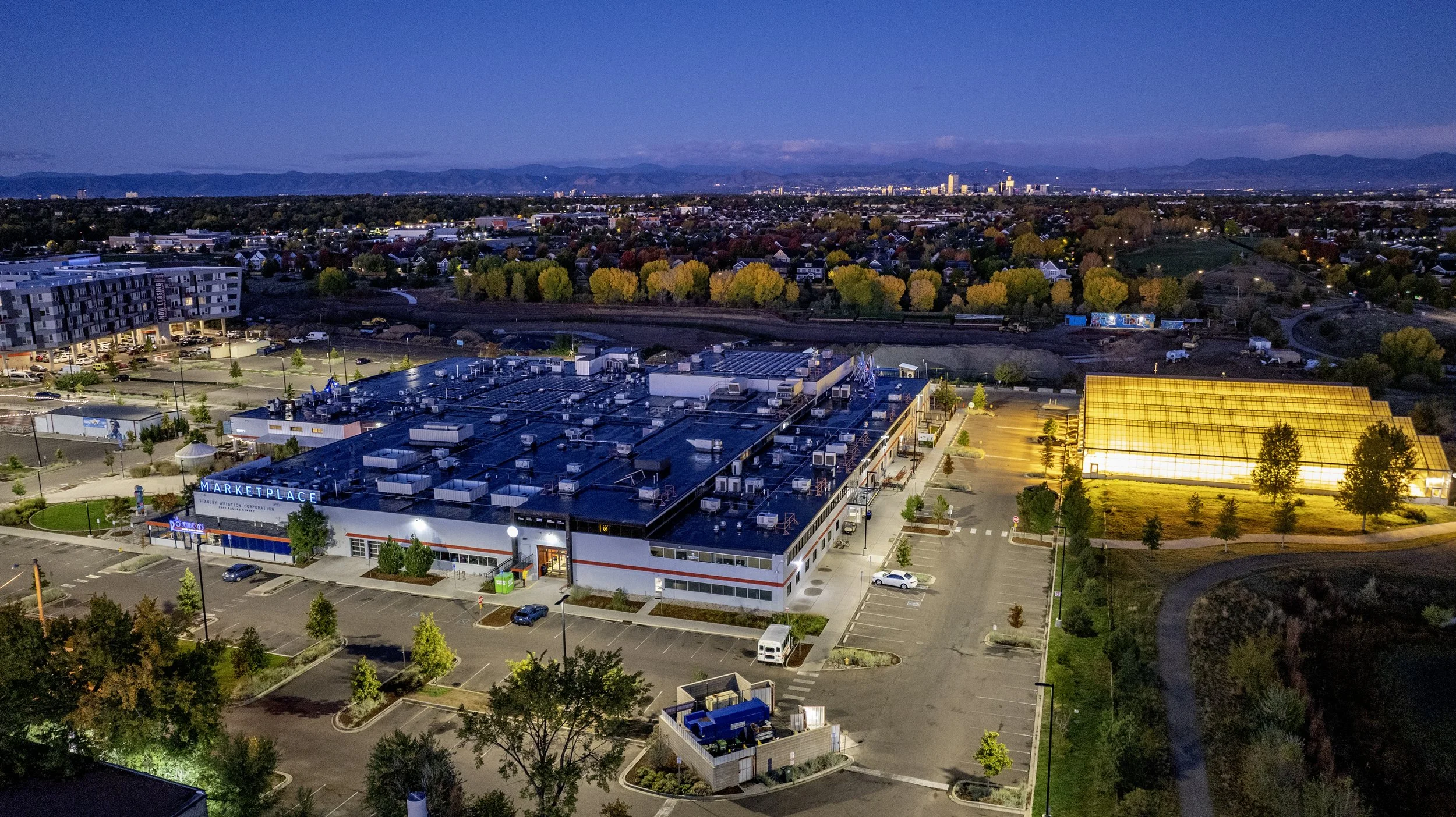An aerial view of a shopping mall and parking lot at dusk, with a cityscape and mountains in the background, and trees lining the area.