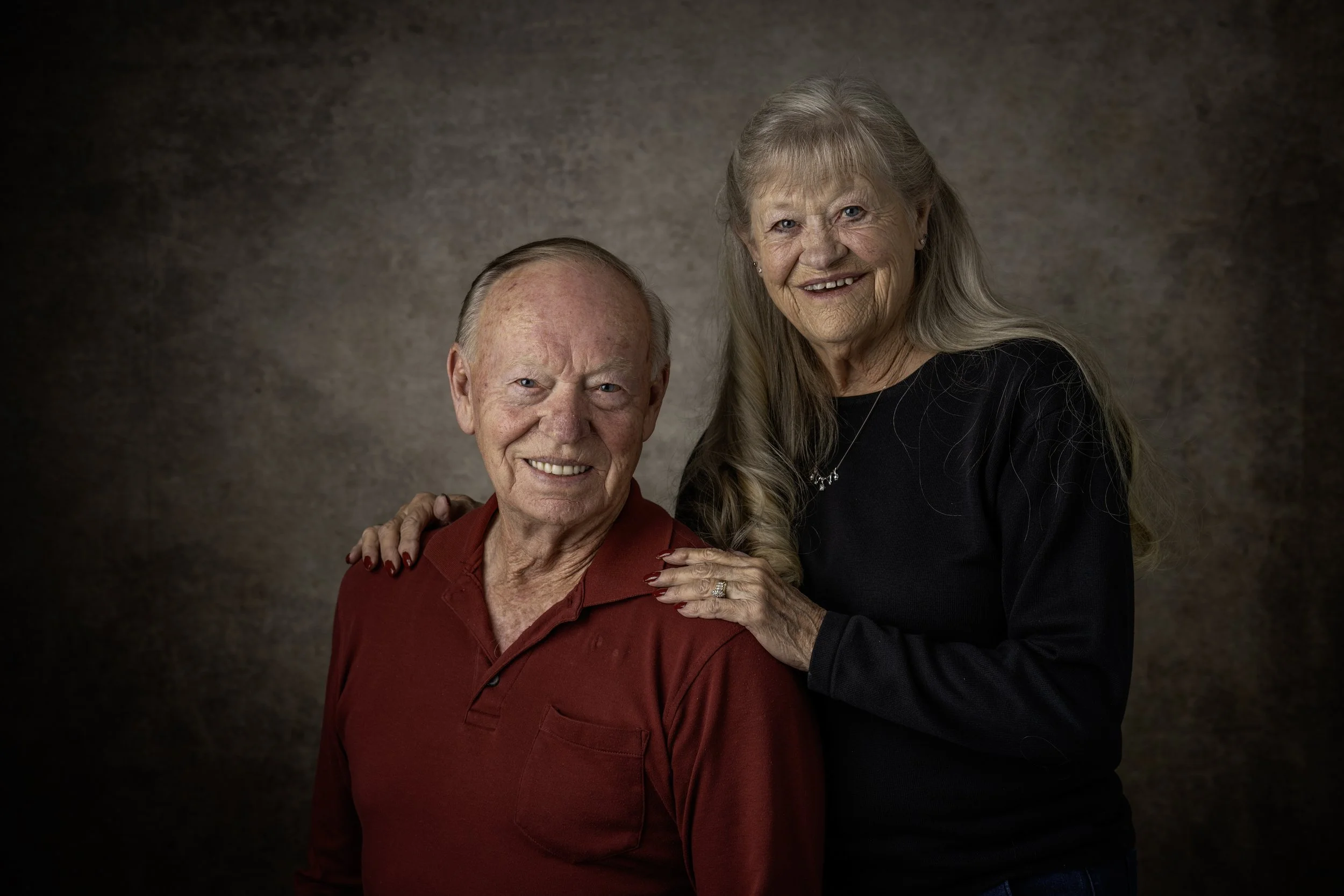 An elderly man and woman smiling at the camera, with the woman resting her hand on the man's shoulder, against a neutral brown background.