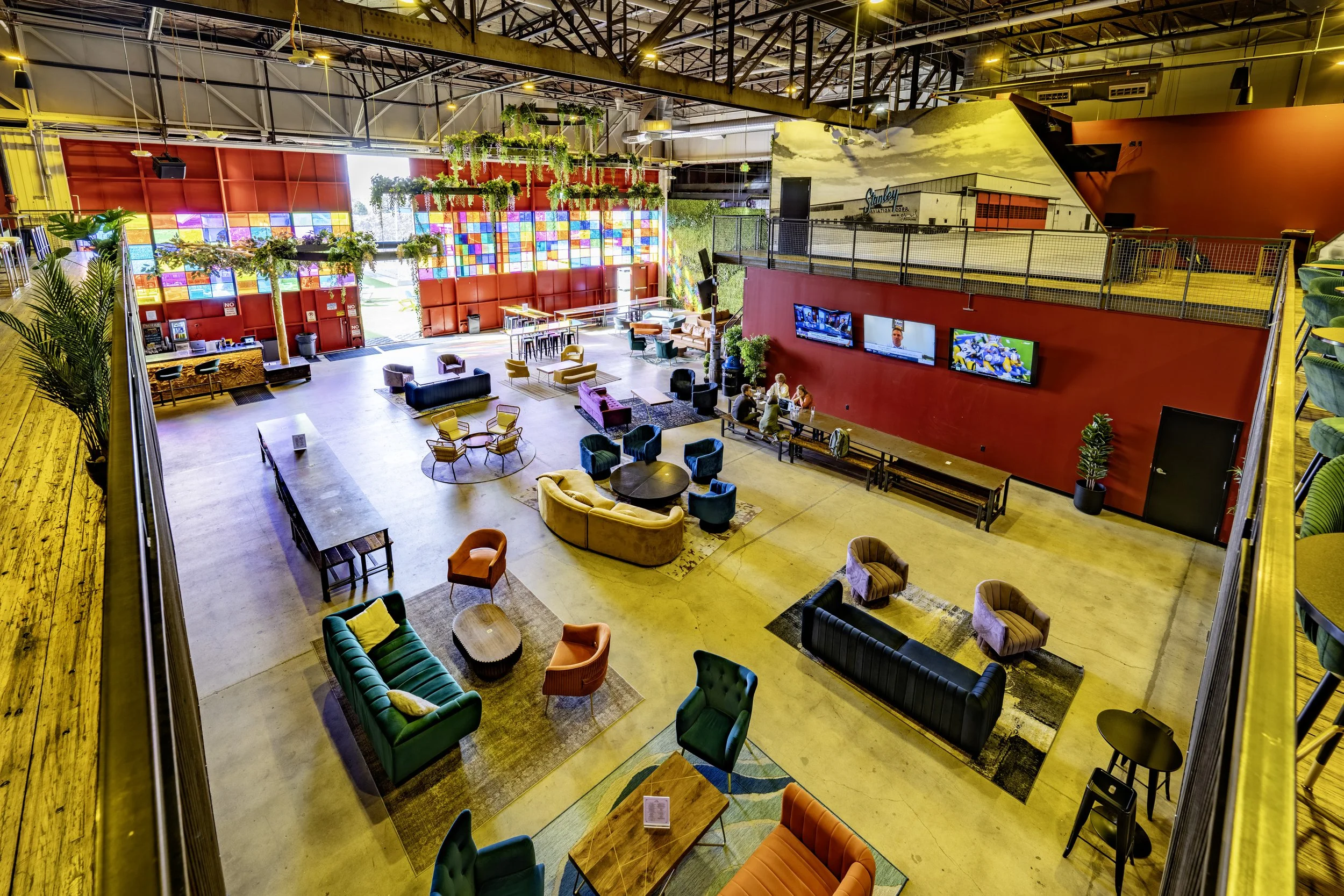 Large indoor lounge area with colorful furniture, including sofas and chairs, a high ceiling, multiple TV screens on a red wall, and large windows allowing natural light. Decor includes hanging plants and potted plants.