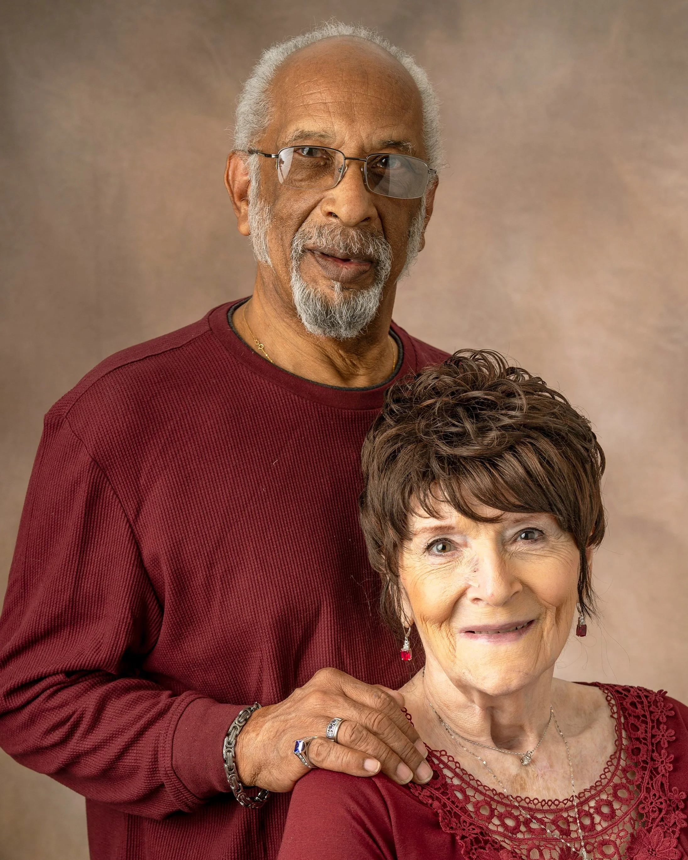 An elderly couple poses together, both wearing red clothing. The man has gray hair, a beard, glasses, and is standing behind the woman, resting his hand on her shoulder. The woman has short, wavy brown hair and is smiling, wearing earrings and neckla