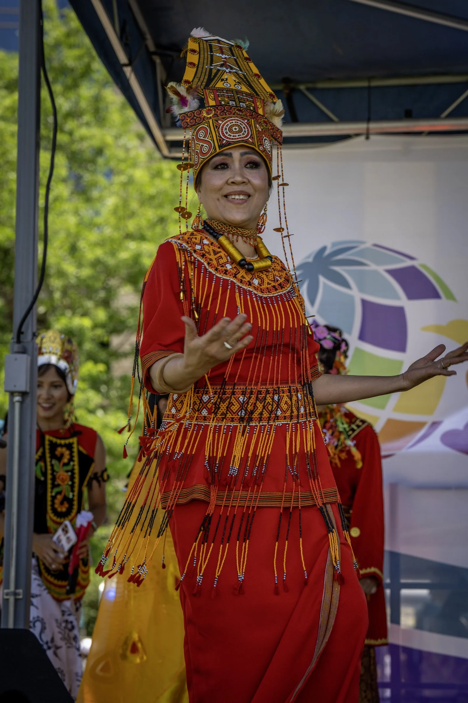 A woman dressed in traditional colorful native attire with intricate beadwork and a tall, decorated headdress, performing or speaking at an outdoor event.