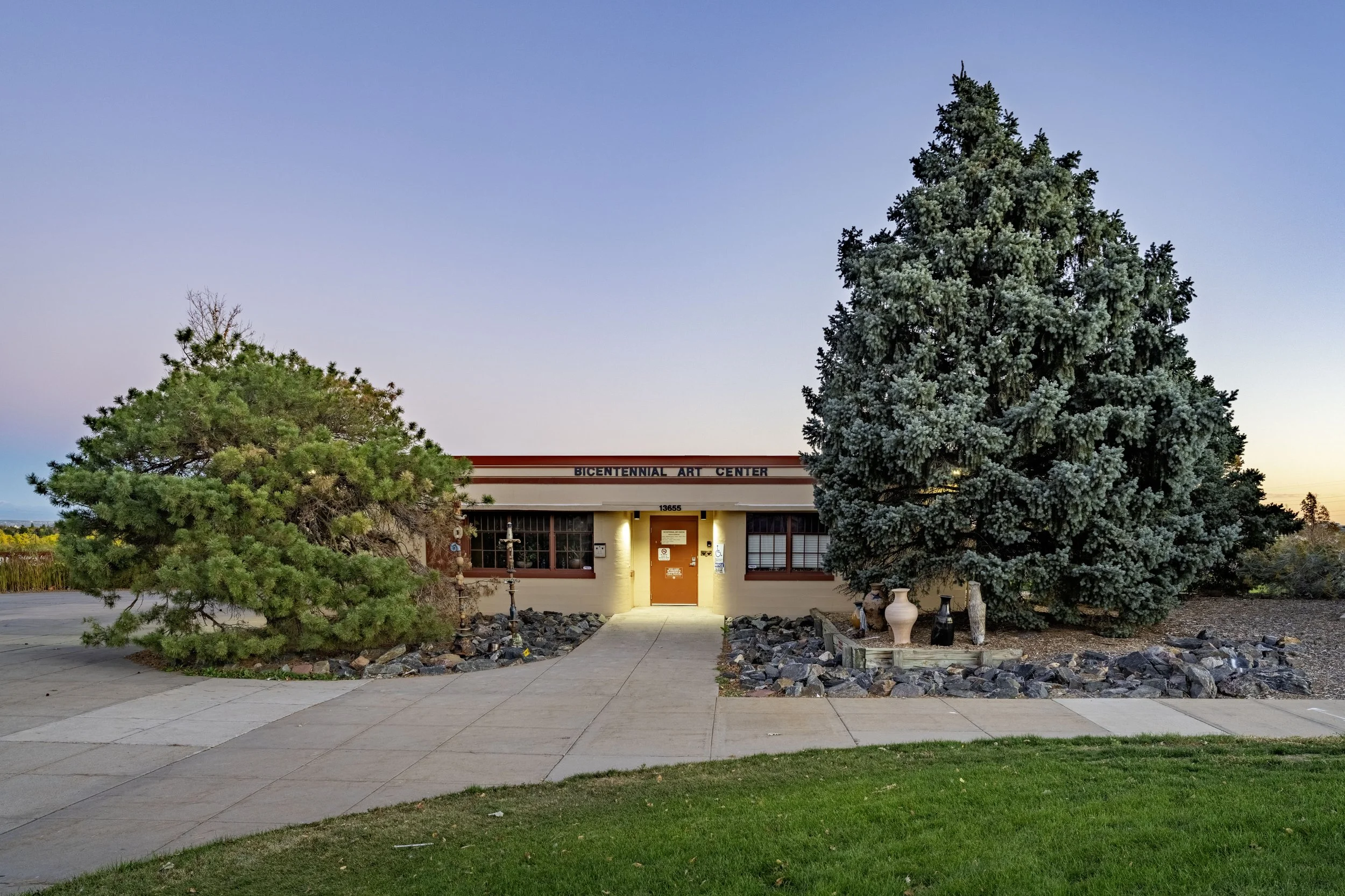 Front view of the Bicentennial Art Center building with two large evergreen trees on each side and a sidewalk leading to the entrance, during sunset.