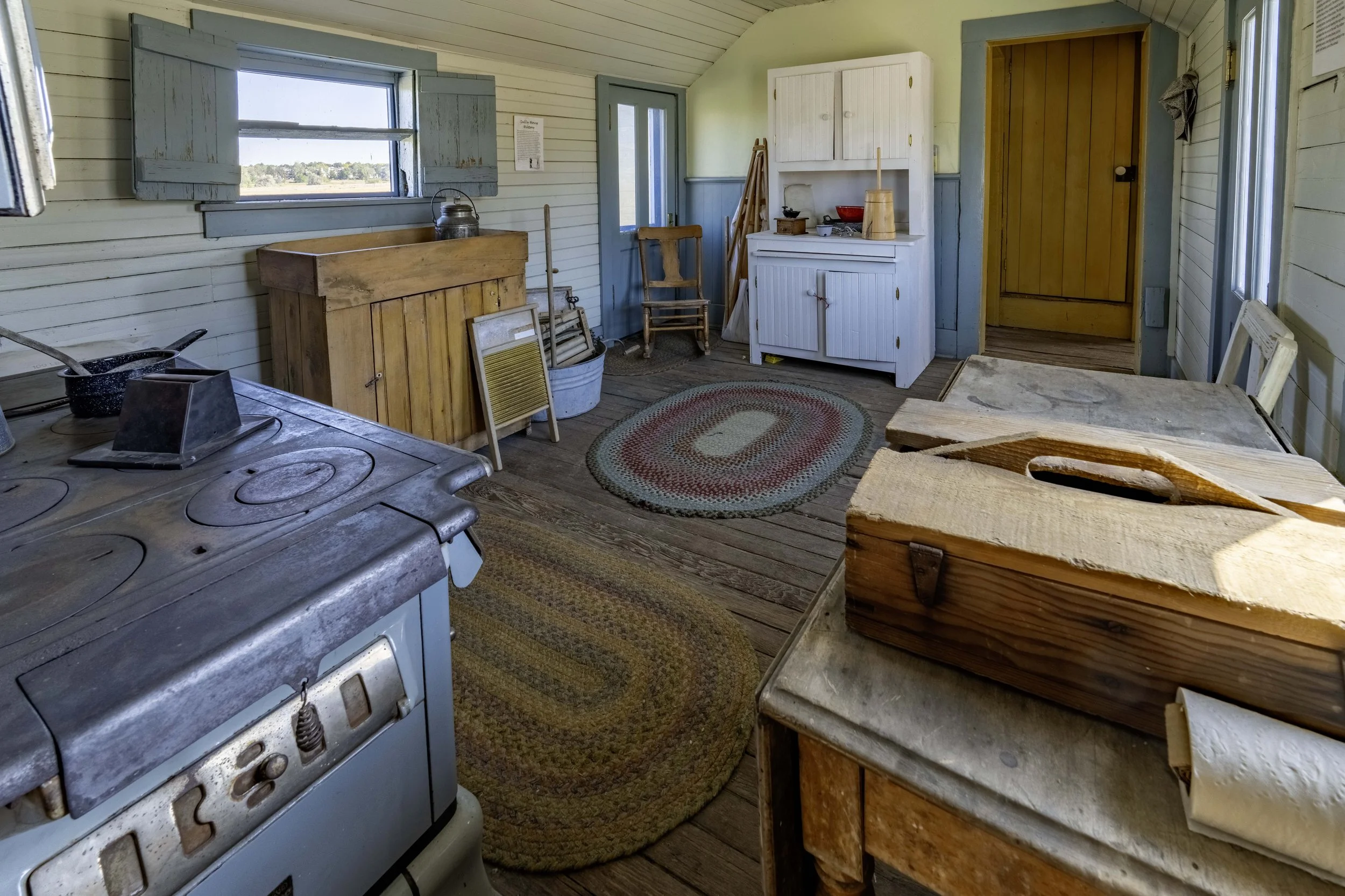 Interior of a rustic, vintage kitchen with wooden floors and walls, featuring an old stove, a wooden table, woven rugs, storage cabinets, and various kitchen utensils and furniture.