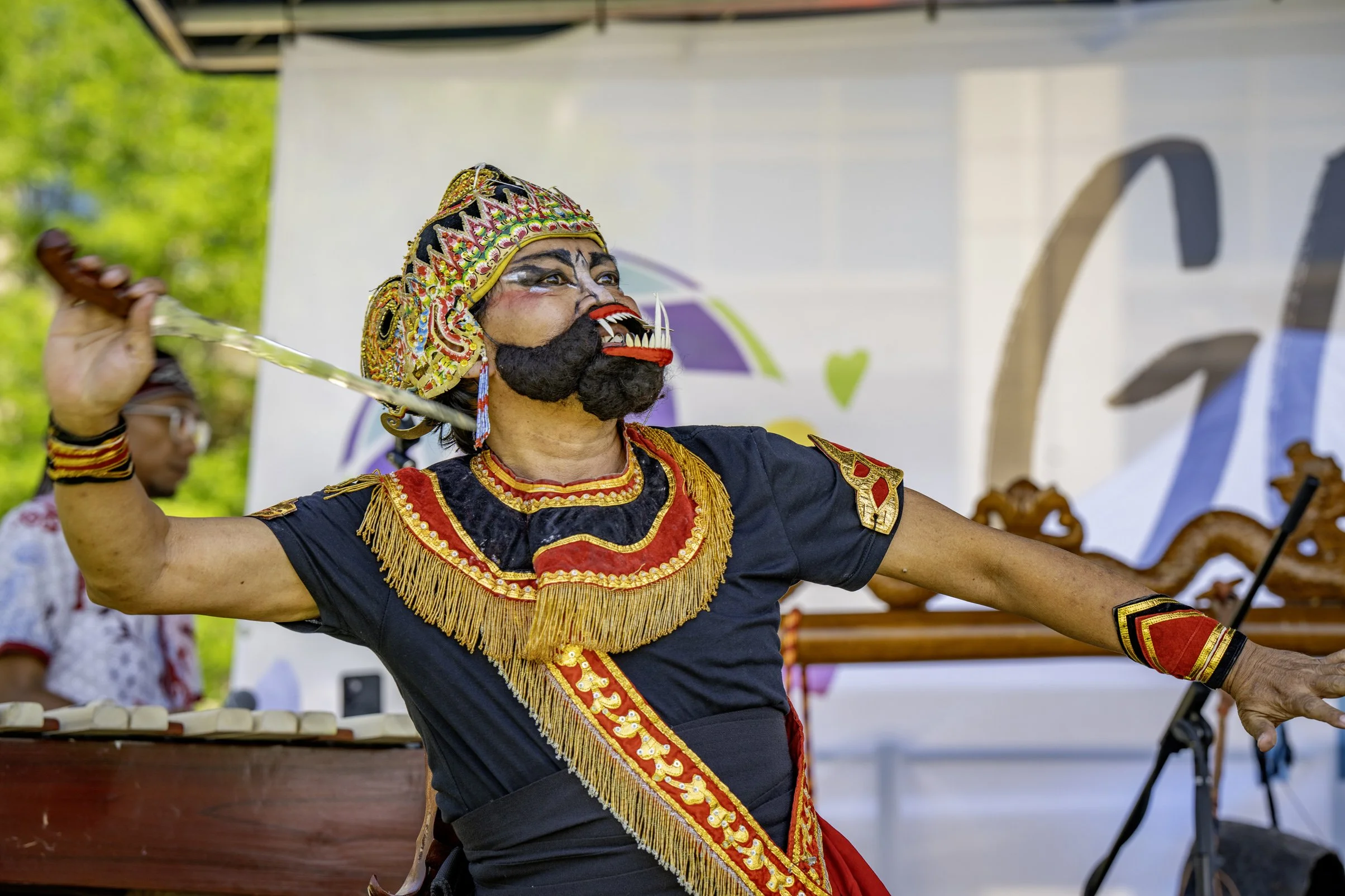 Cultural performer in traditional costume with mask, holding a dagger, participating in a cultural or dance event outdoors.