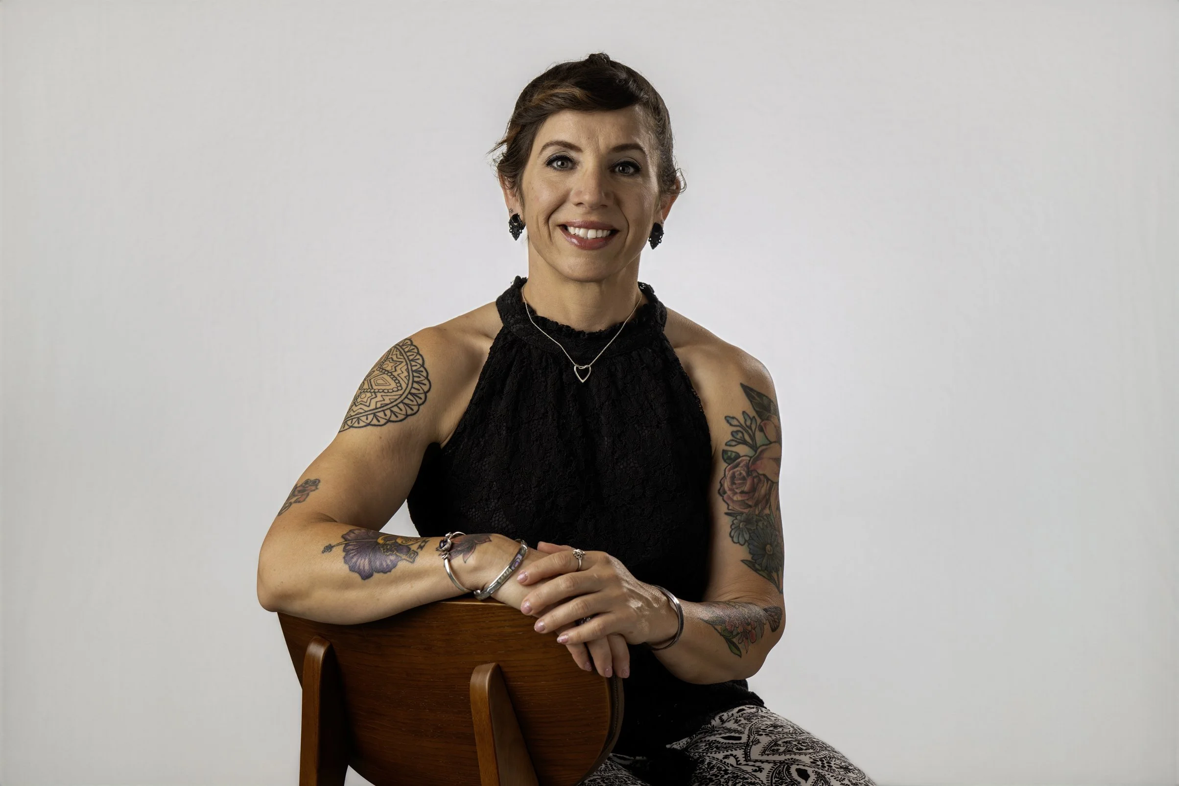 A smiling woman with short brown hair and tattoos on her arms, wearing a black sleeveless top, jewelry, and patterned pants, sitting on a wooden chair against a plain white background.