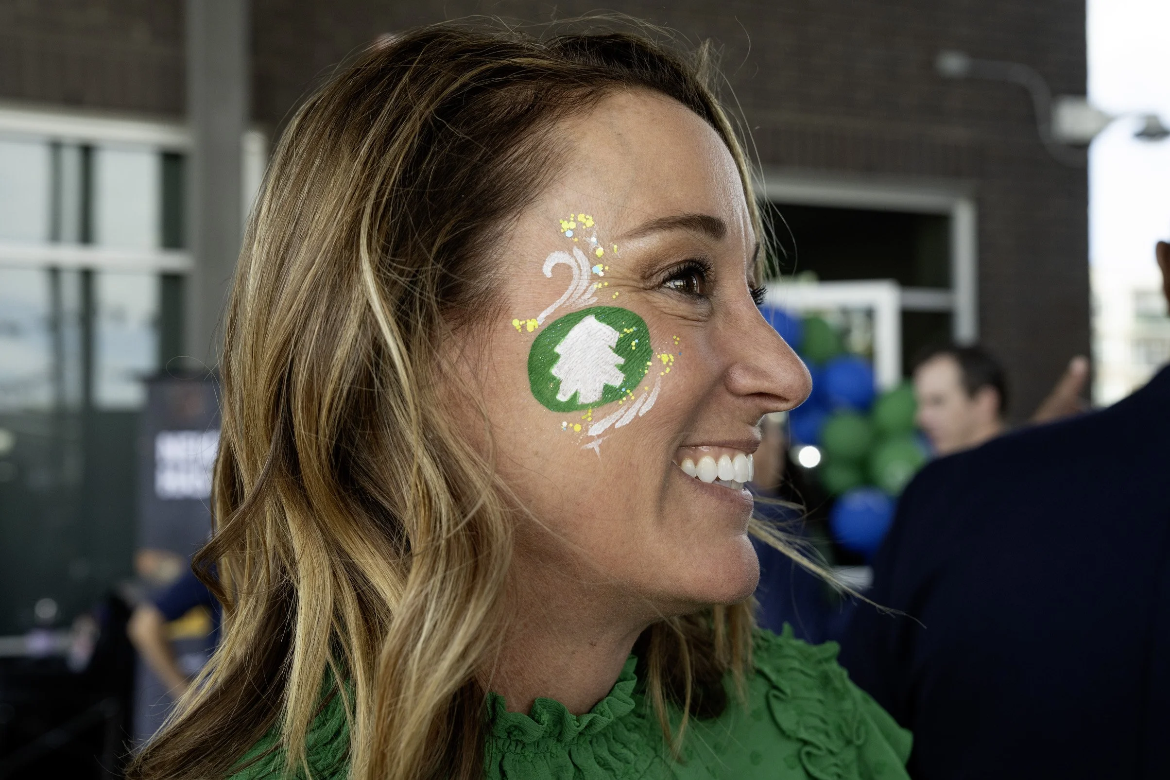 A woman with face paint celebrating a 2nd anniversary, wearing a green top, at an outdoor event with balloons and other people in the background.
