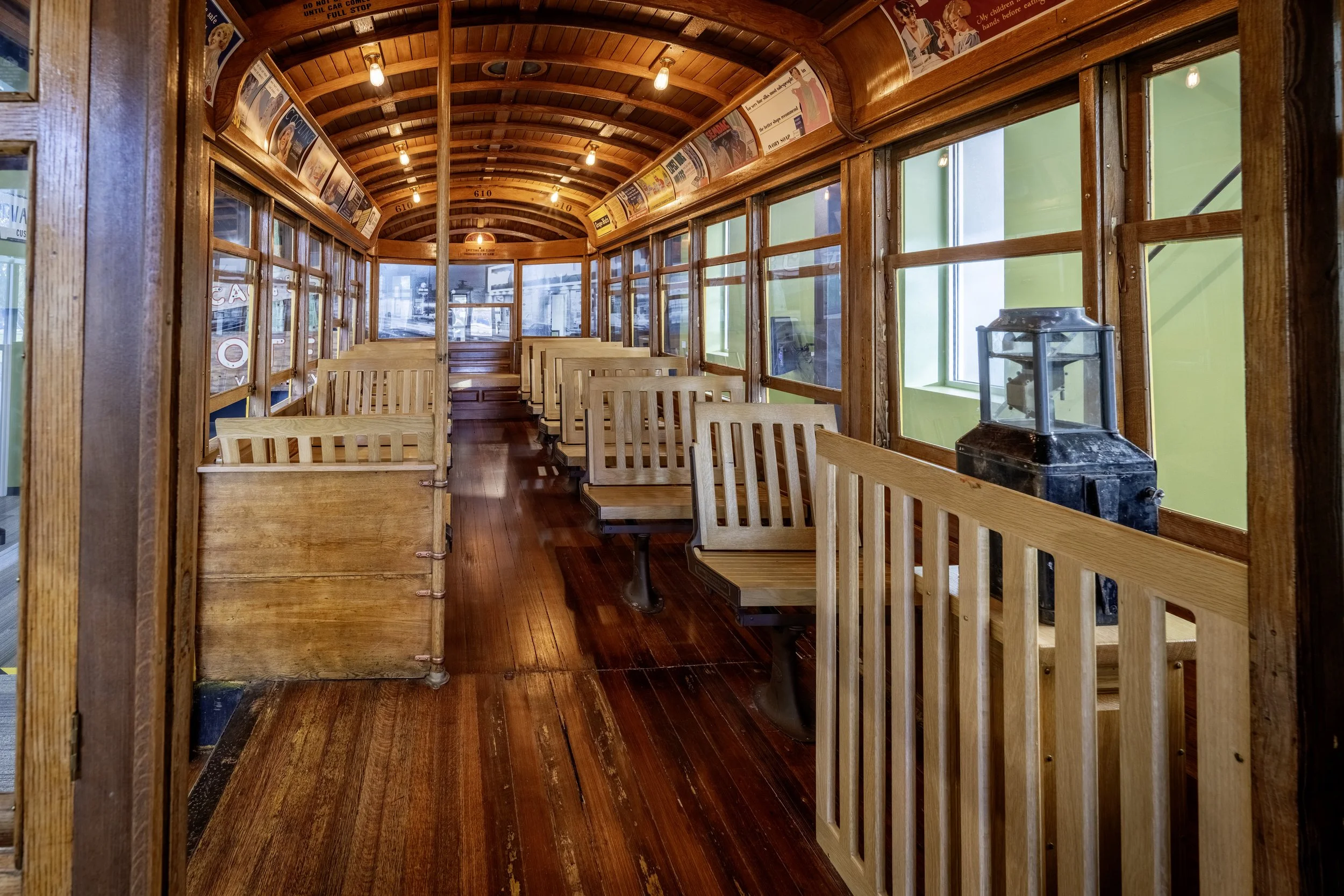 Interior of a vintage train car with wooden benches and floors, green-tinted windows, and a lantern on a stand near the window.