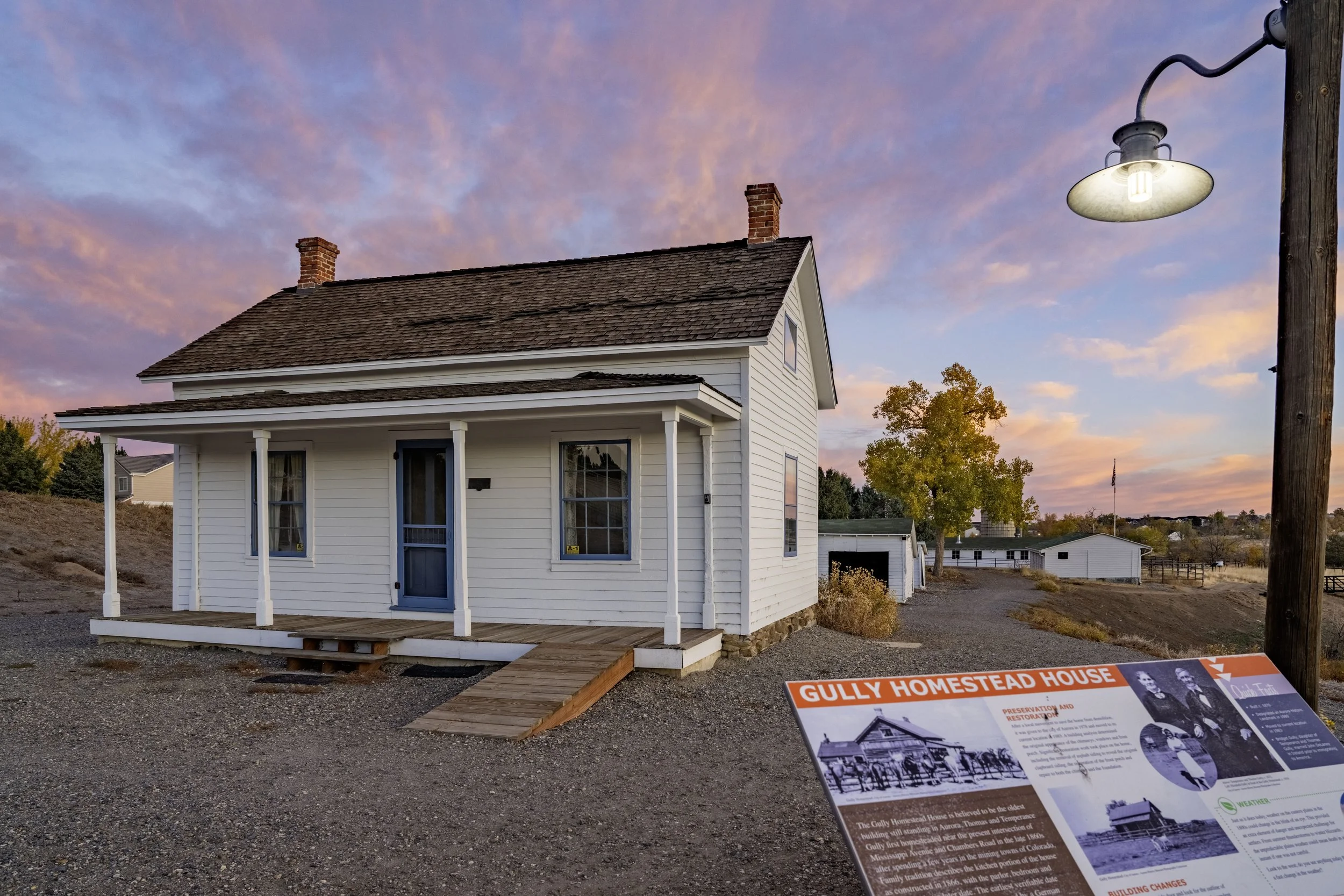 A historic white Gully Homestead House with a porch and ramp, situated outdoors at sunset with a pink and purple sky. There is a sign at the front providing information about its preservation and restoration.