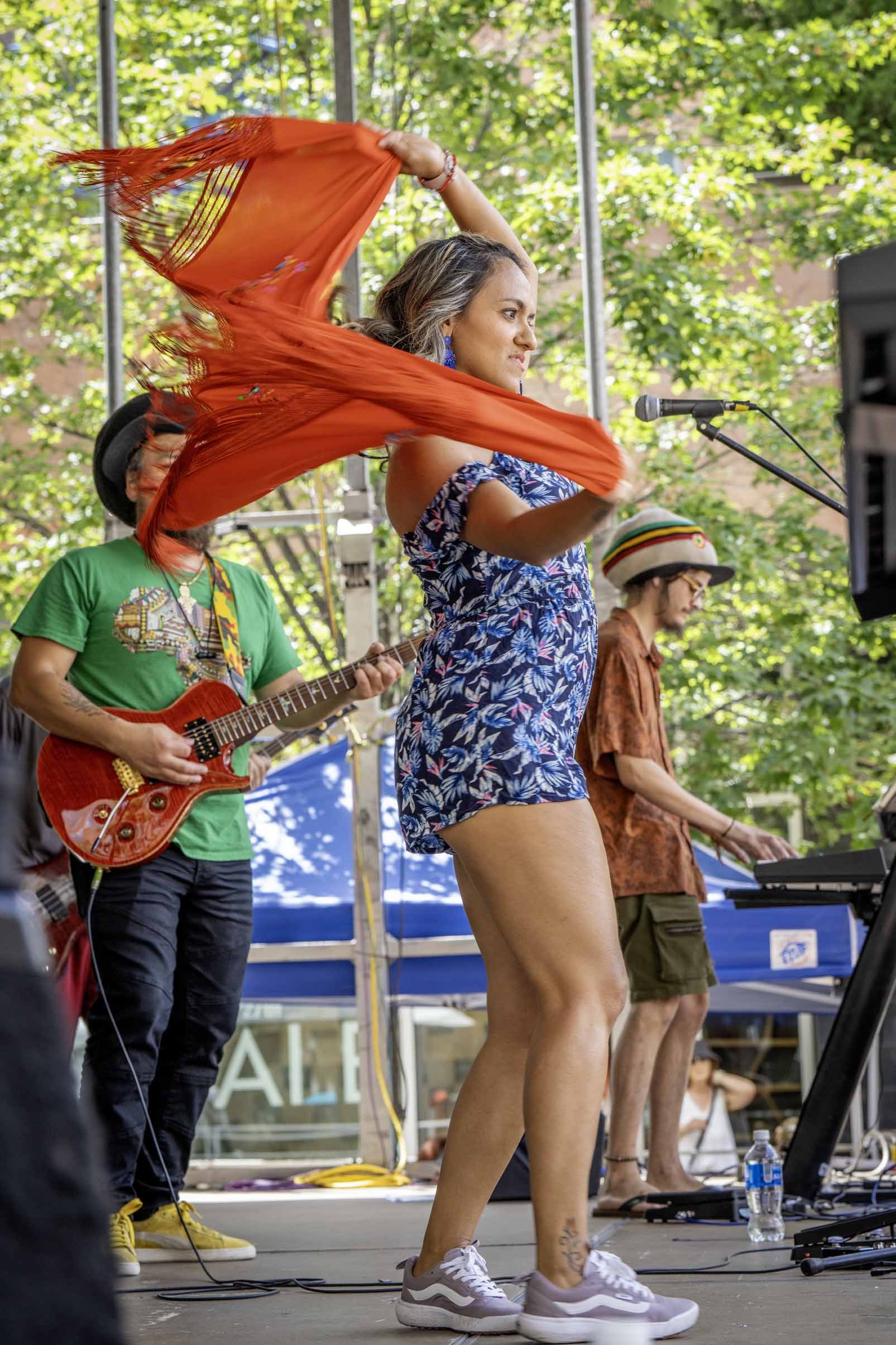 A woman dancing on stage with a red scarf, flanked by musicians playing guitars and keyboards, outdoors under green trees.