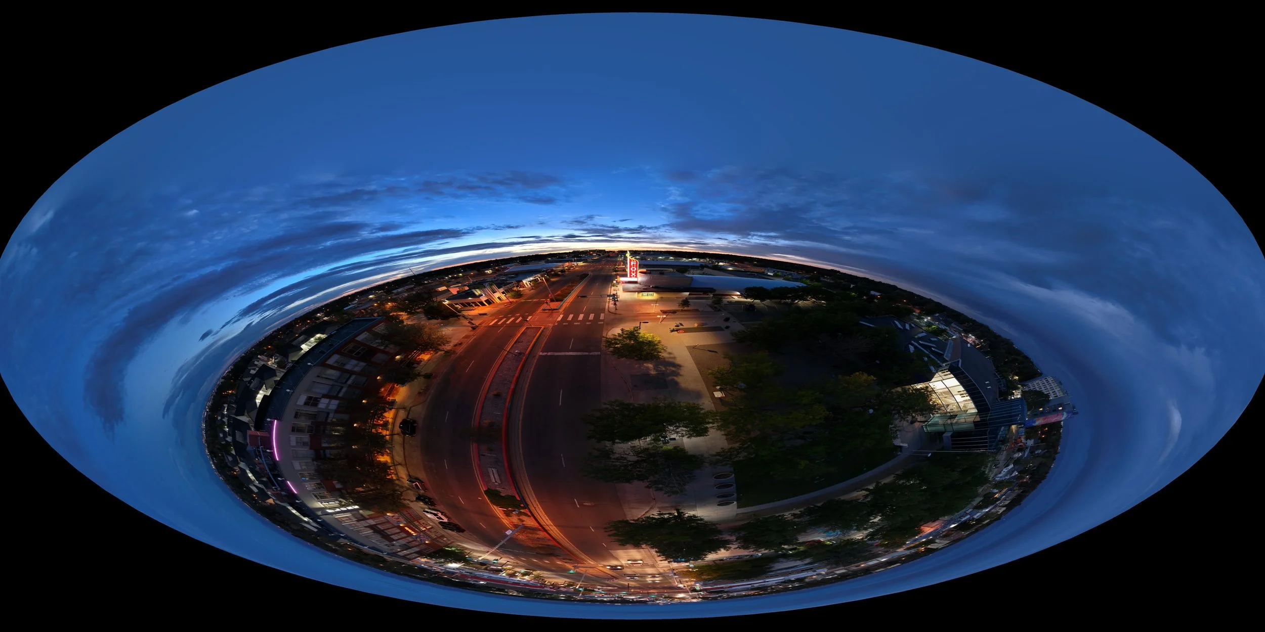 A panoramic, fisheye view of a cityscape during dusk with blue sky, clouds, trees, buildings, and illuminated streets.