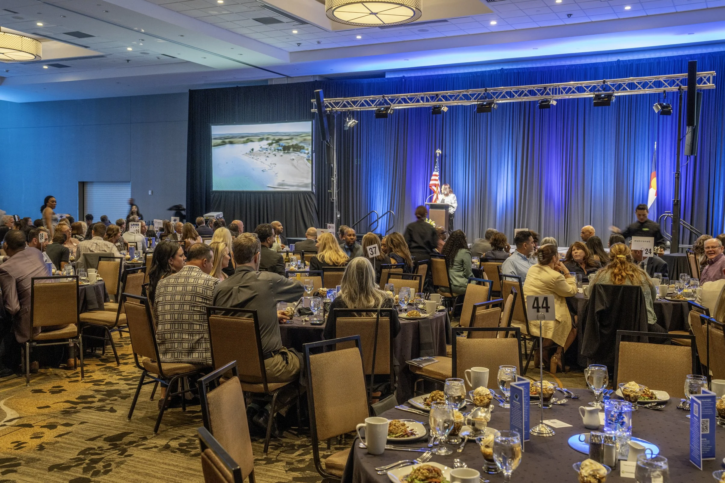 A large conference room filled with round tables and many attendees sitting and listening to a speaker at a podium on stage. The stage has blue curtains, an American flag, a screen showing a beach scene, and a Colorado state flag.