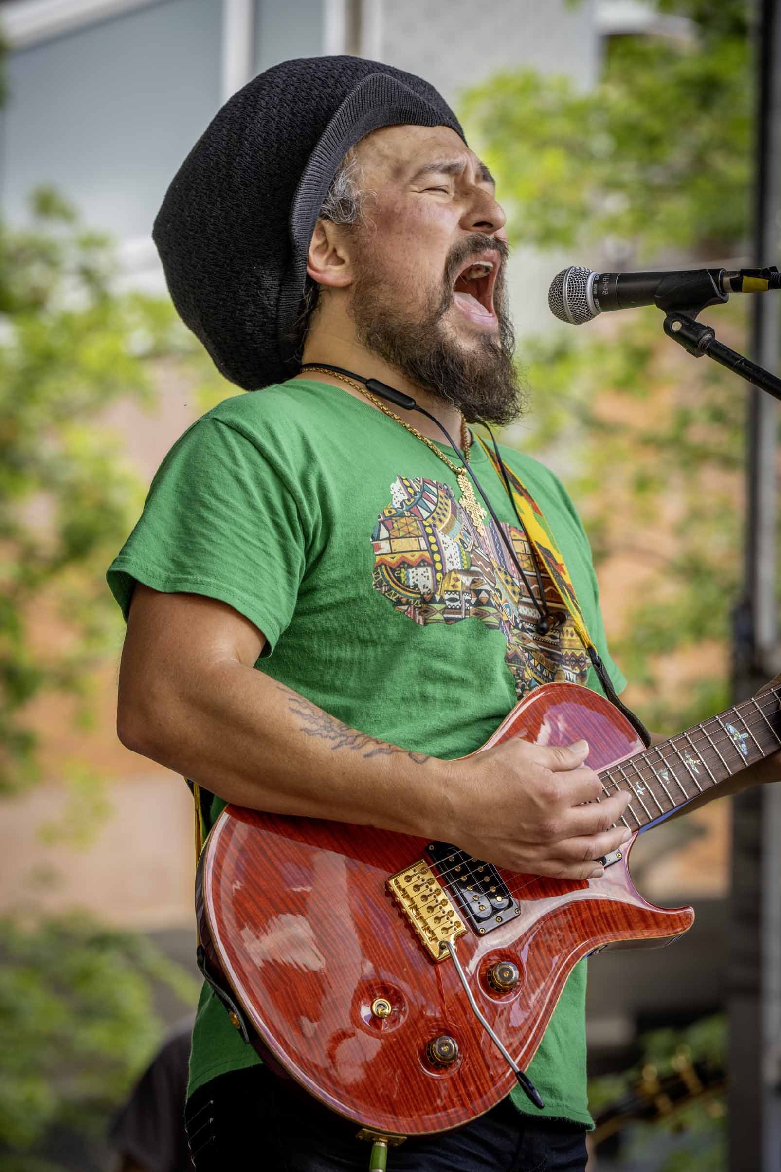 A man passionately singing into a microphone while playing an electric guitar outdoors. He has a beard, wears a black hat, a green t-shirt, and necklaces, with a colorful tattoo on his arm.