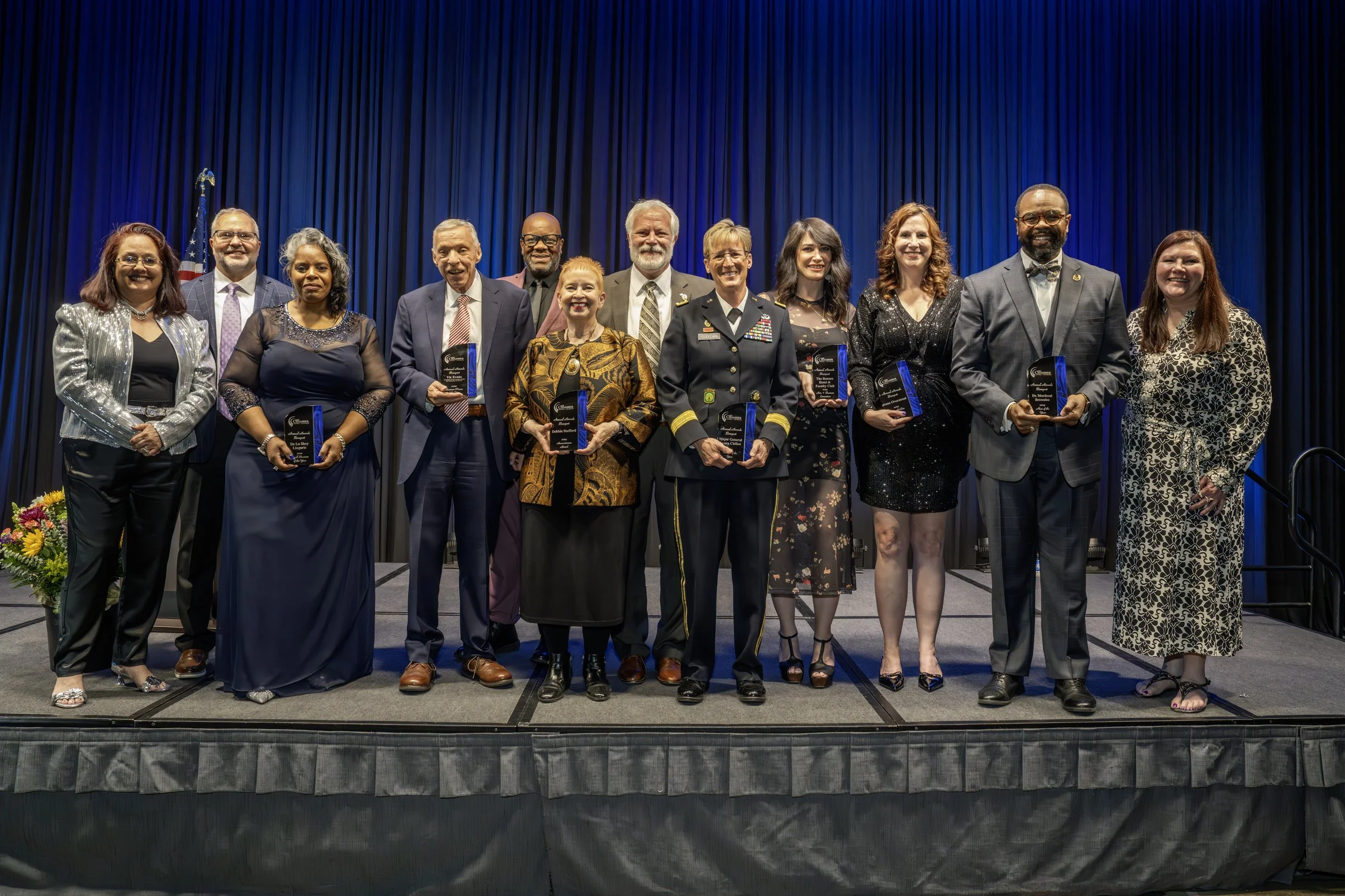 Group of 13 diverse people standing on stage holding awards, with a dark curtain background and American flag on the left.