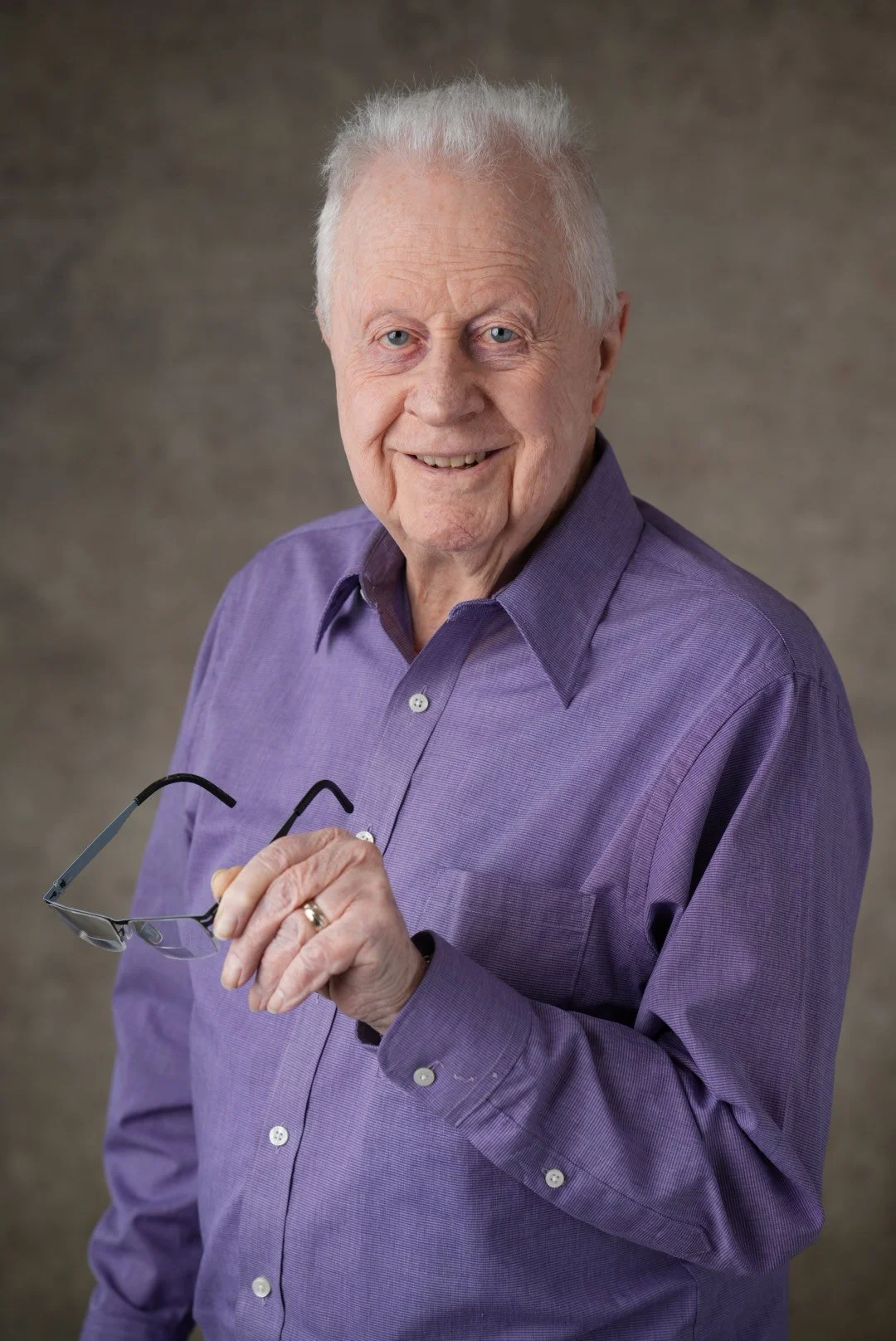 An elderly man with white hair and blue eyes is smiling and holding a pair of glasses in his right hand. He is wearing a purple button-down shirt and standing against a plain, textured background.