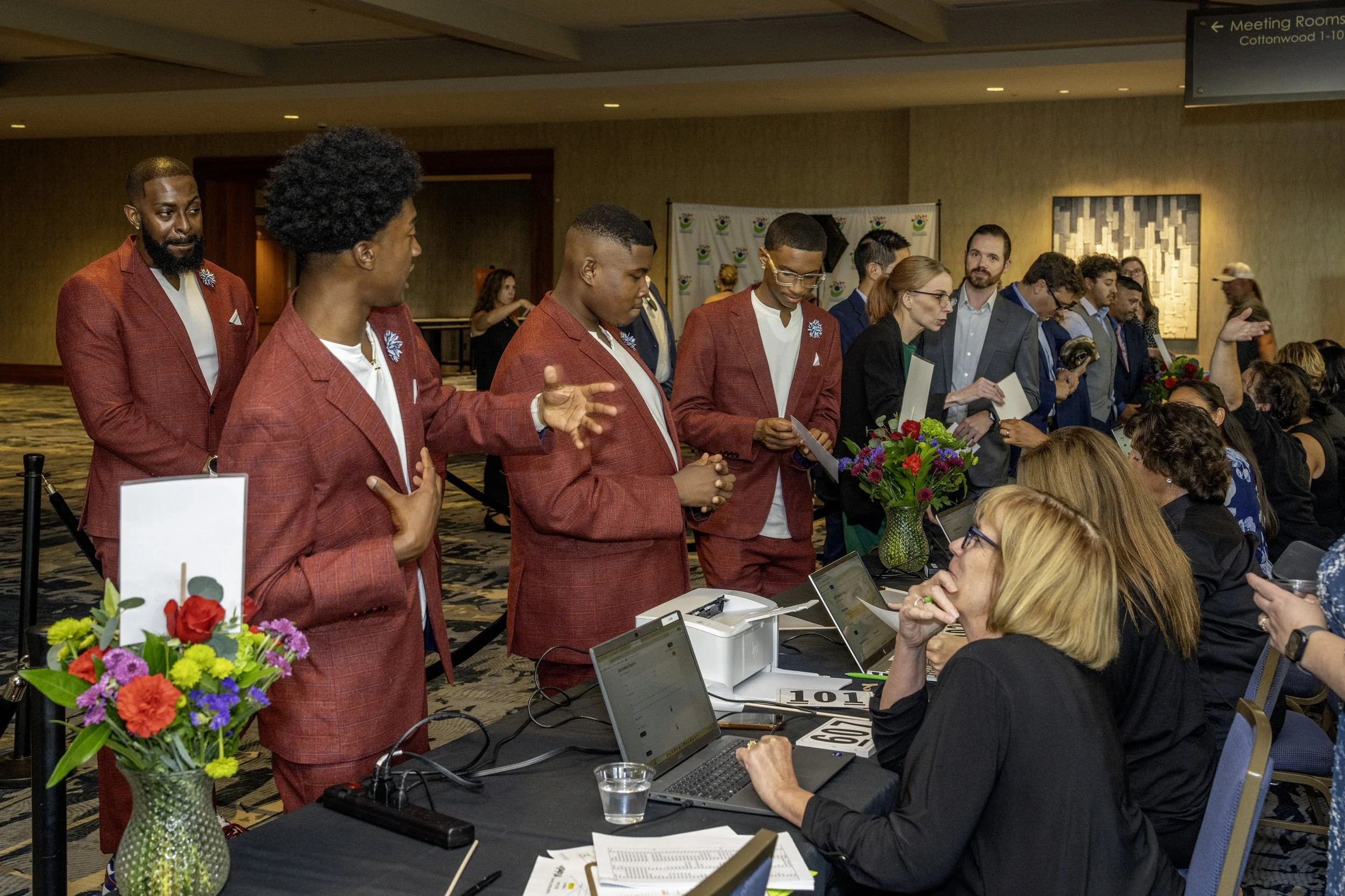 A group of young people in red suits standing at a registration or check-in table at a conference or event, talking to event staff, with laptops and flower arrangements on the table.