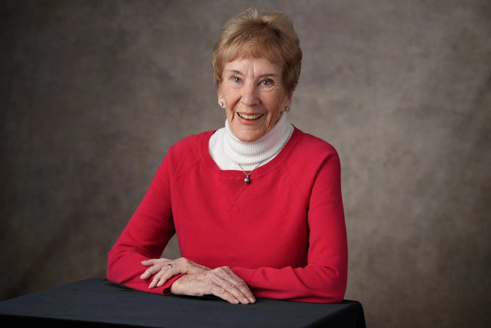 An elderly woman with short, light brown hair, wearing a red sweater over a white turtleneck, smiling at the camera while seated at a black table against a neutral background.