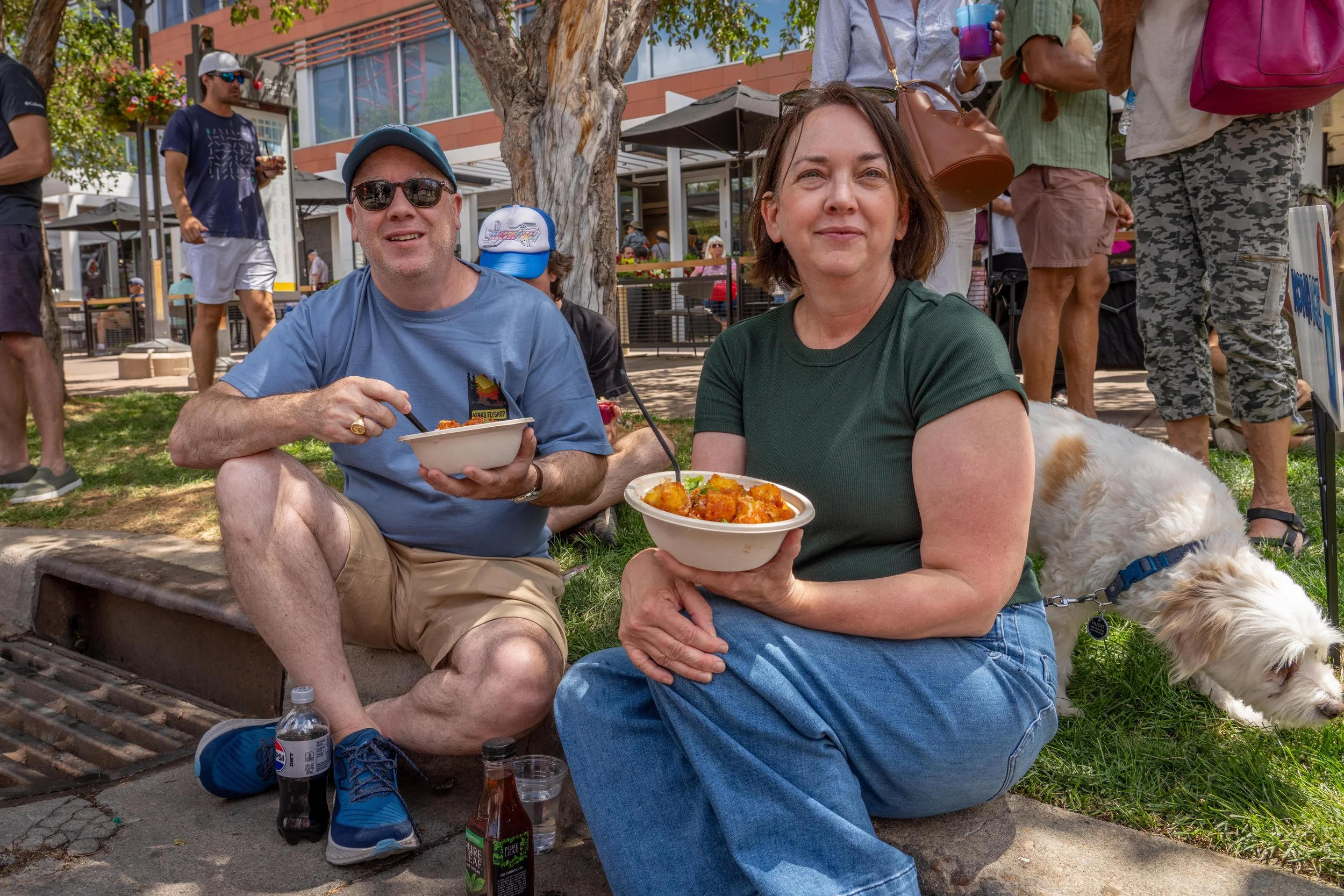 Two people sitting on the grass eating food from bowls, with a dog nearby. They are outdoors in a park or plaza with other people walking and standing around.