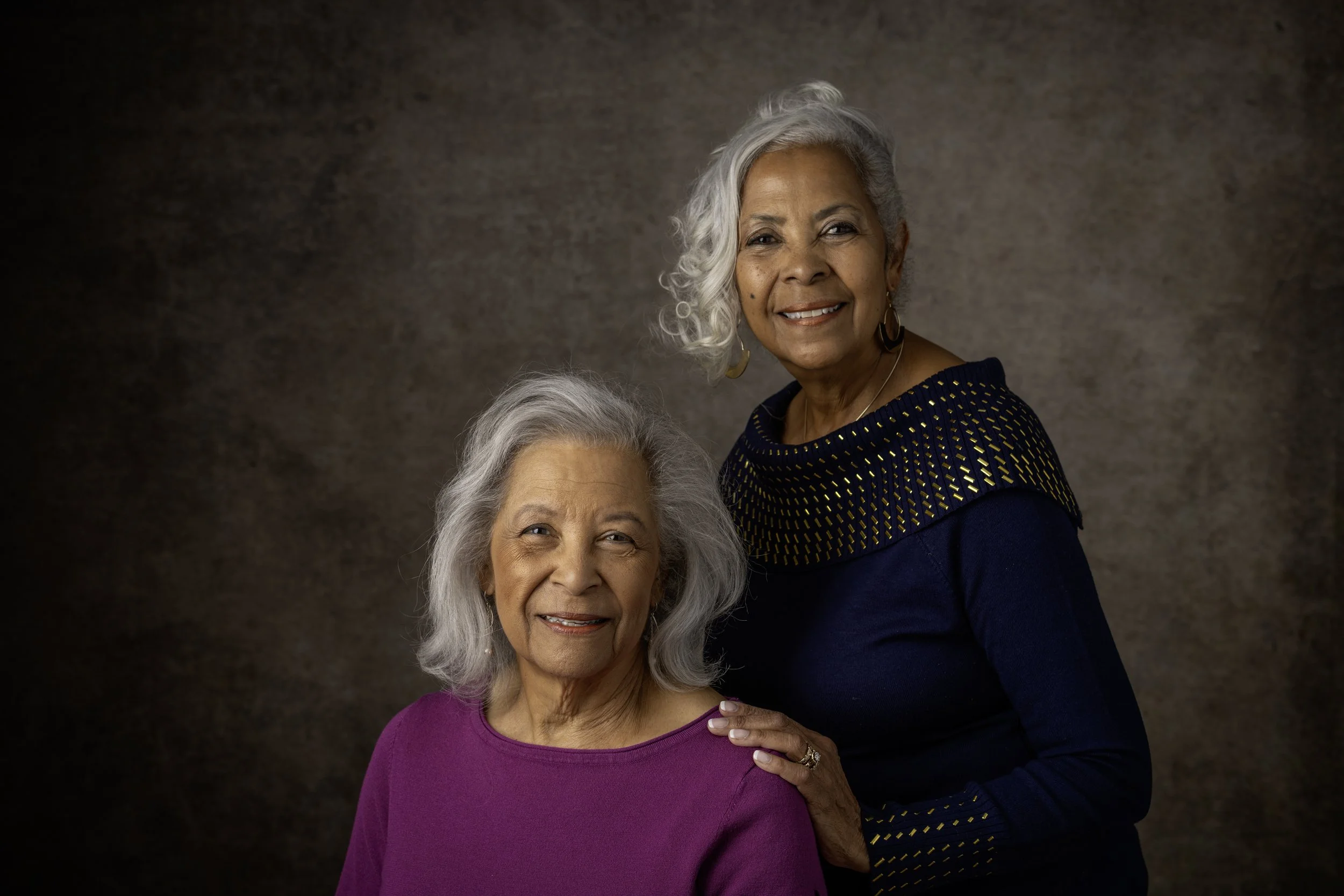 Two elderly women with gray hair posing together against a brown textured background. One is seated wearing a purple top, and the other is standing beside her wearing a dark blue top with yellow accents.