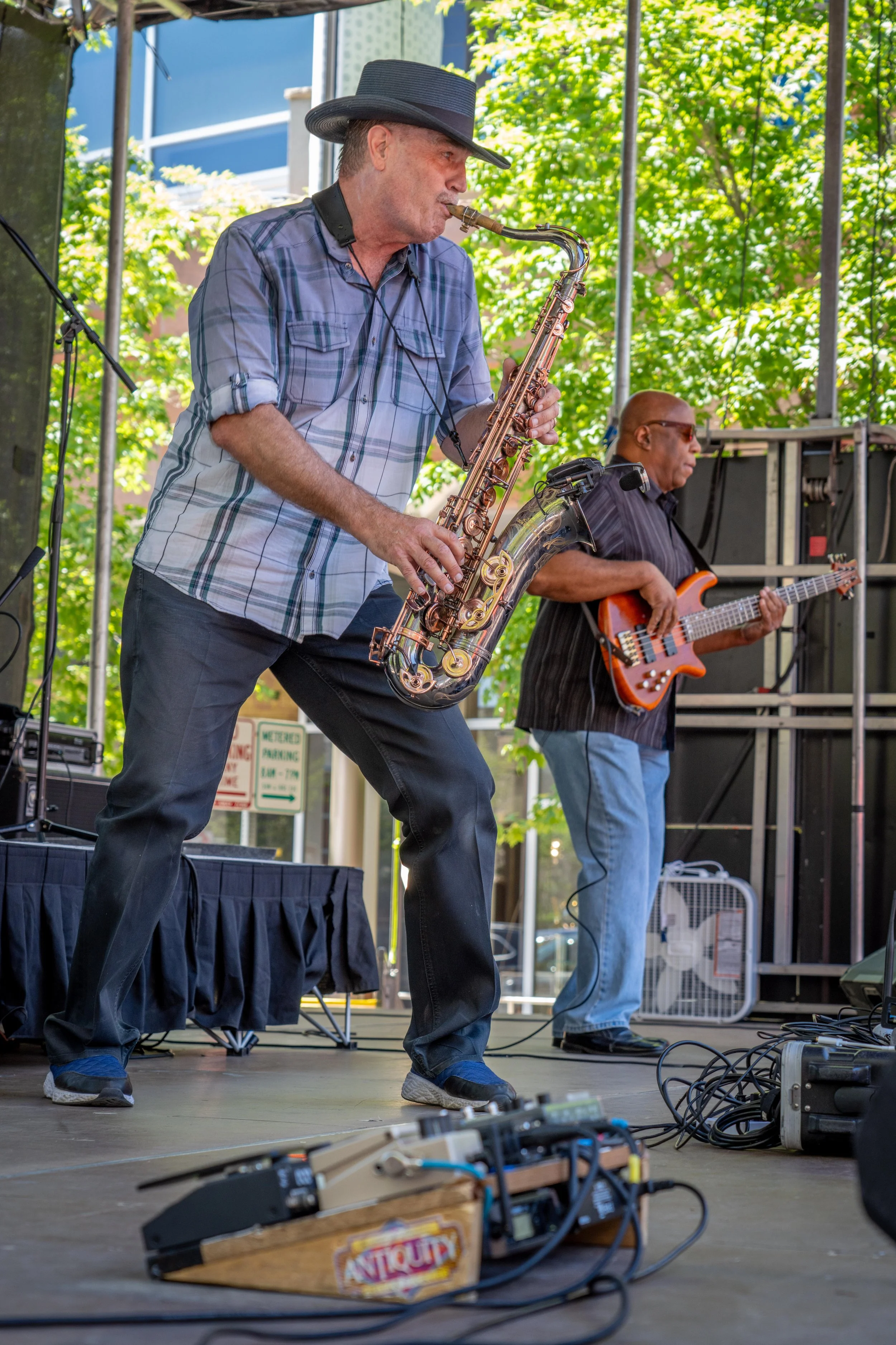 Two musicians performing on an outdoor stage. One playing a saxophone, the other a bass guitar.