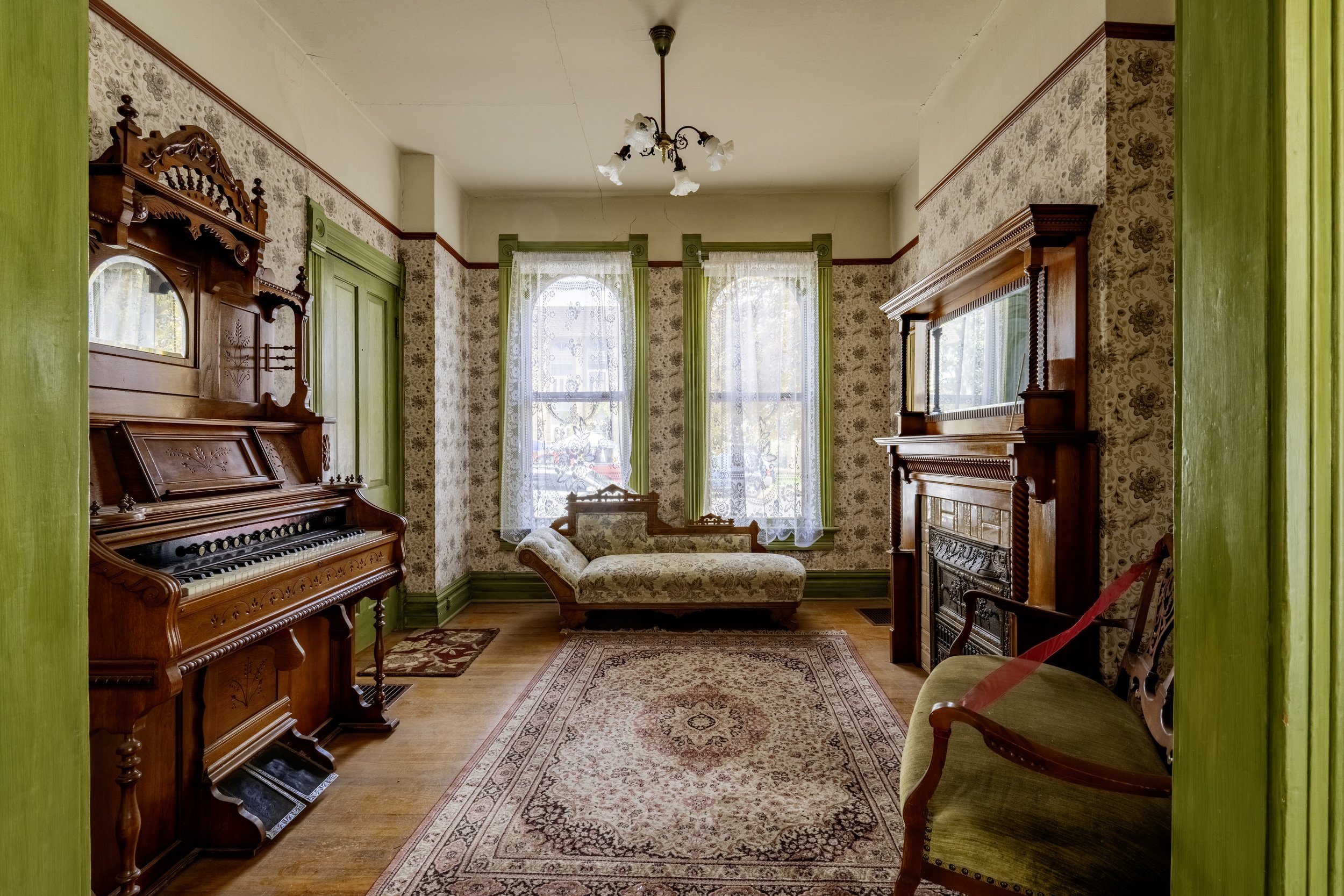 Vintage living room with wooden furniture, decorative wallpaper, and lace curtains, featuring an antique piano, a chaise lounge, and a fireplace.