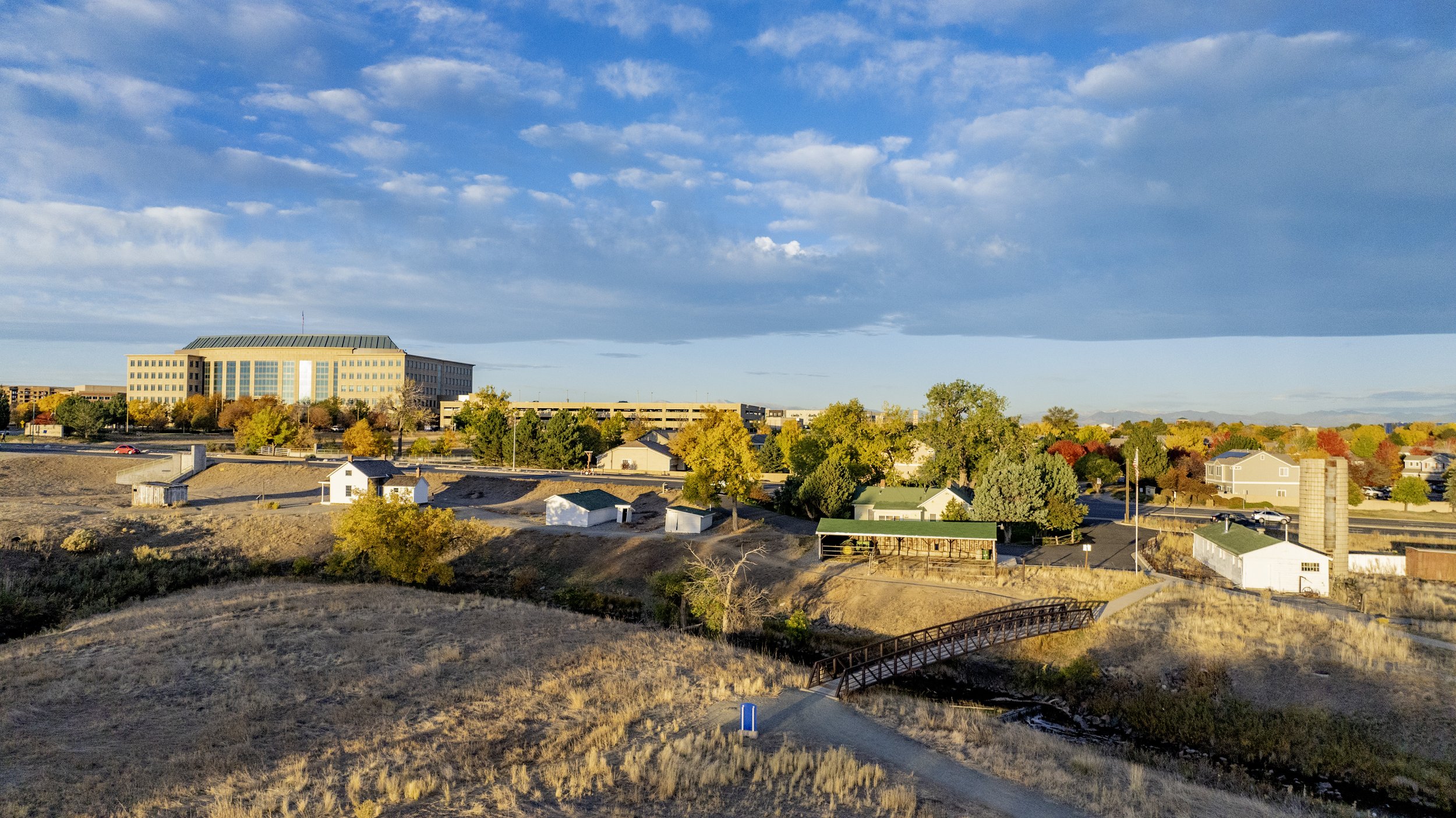 Aerial view of a city landscape with a large modern building, autumn colored trees, small white structures, and a dirt path with a small wooden bridge.