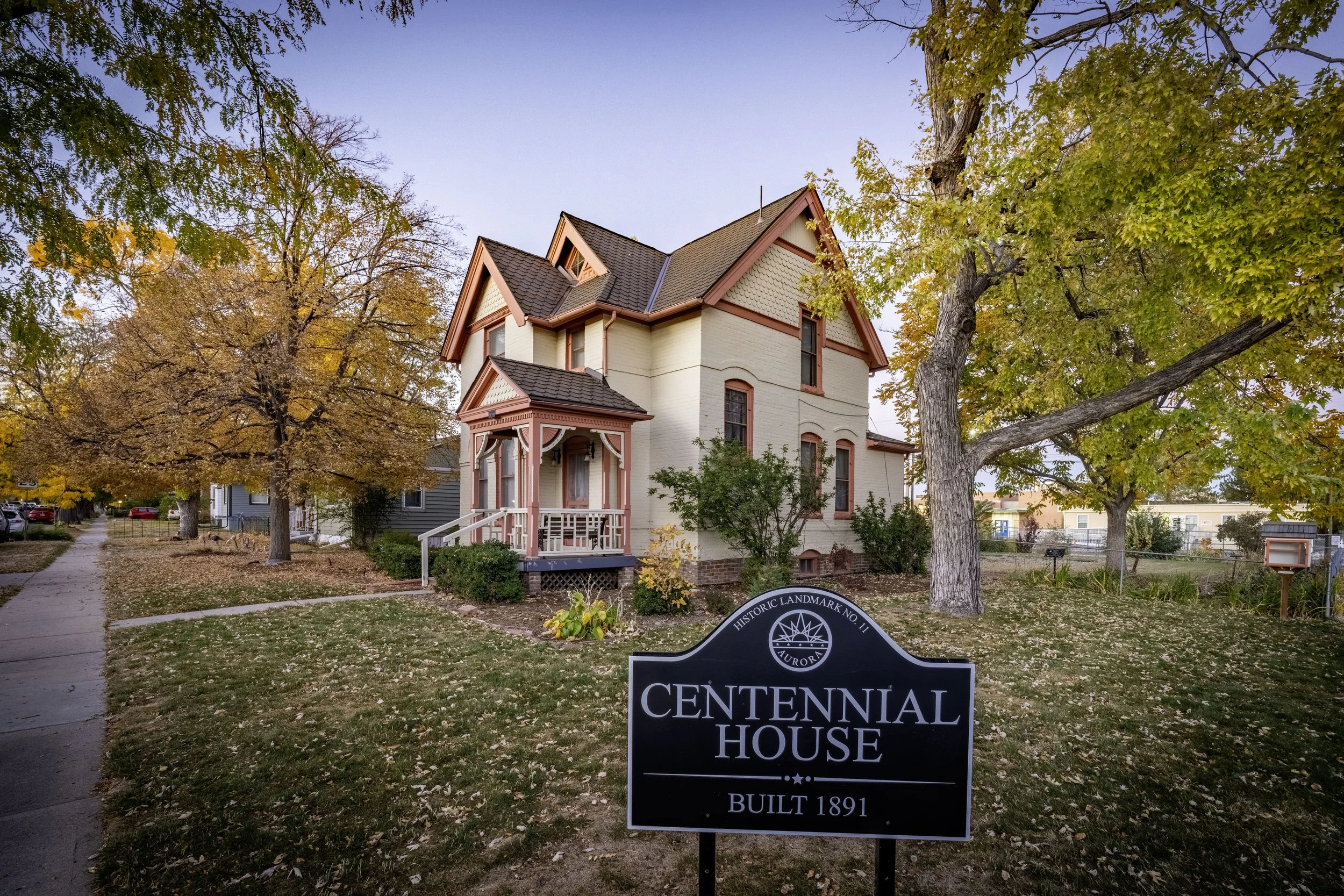 A historic 1891 Victorian house with cream walls, pink trim, and a small porch, surrounded by trees with autumn leaves. A black sign in the front reads "Centennial House, Built 1891."