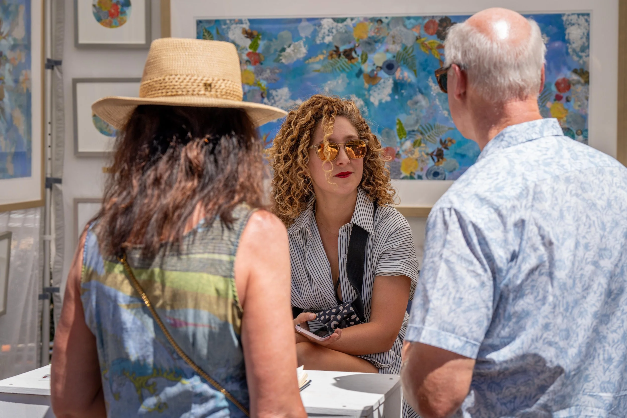 Three people at an art gallery, two women and one man, engaged in conversation. The woman in the middle is wearing sunglasses, a striped shirt, and holding a phone. The woman on the left is wearing a straw hat and patterned tank top. The man on the r