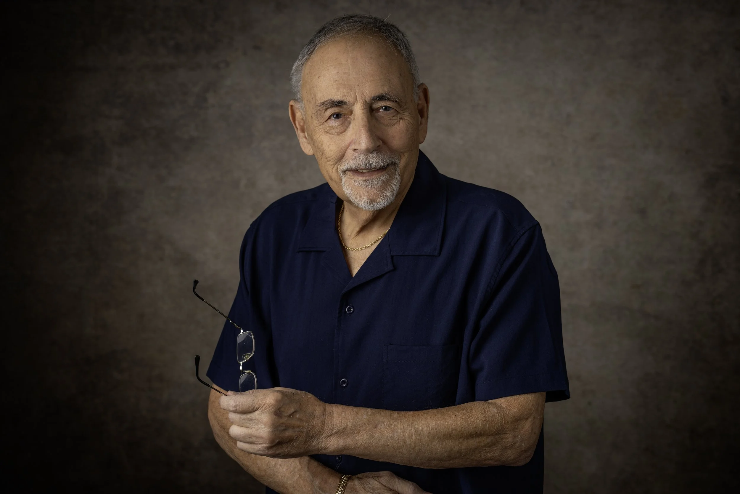 Older man with gray hair and beard wearing a navy blue shirt, holding glasses, smiling, against brown textured background.