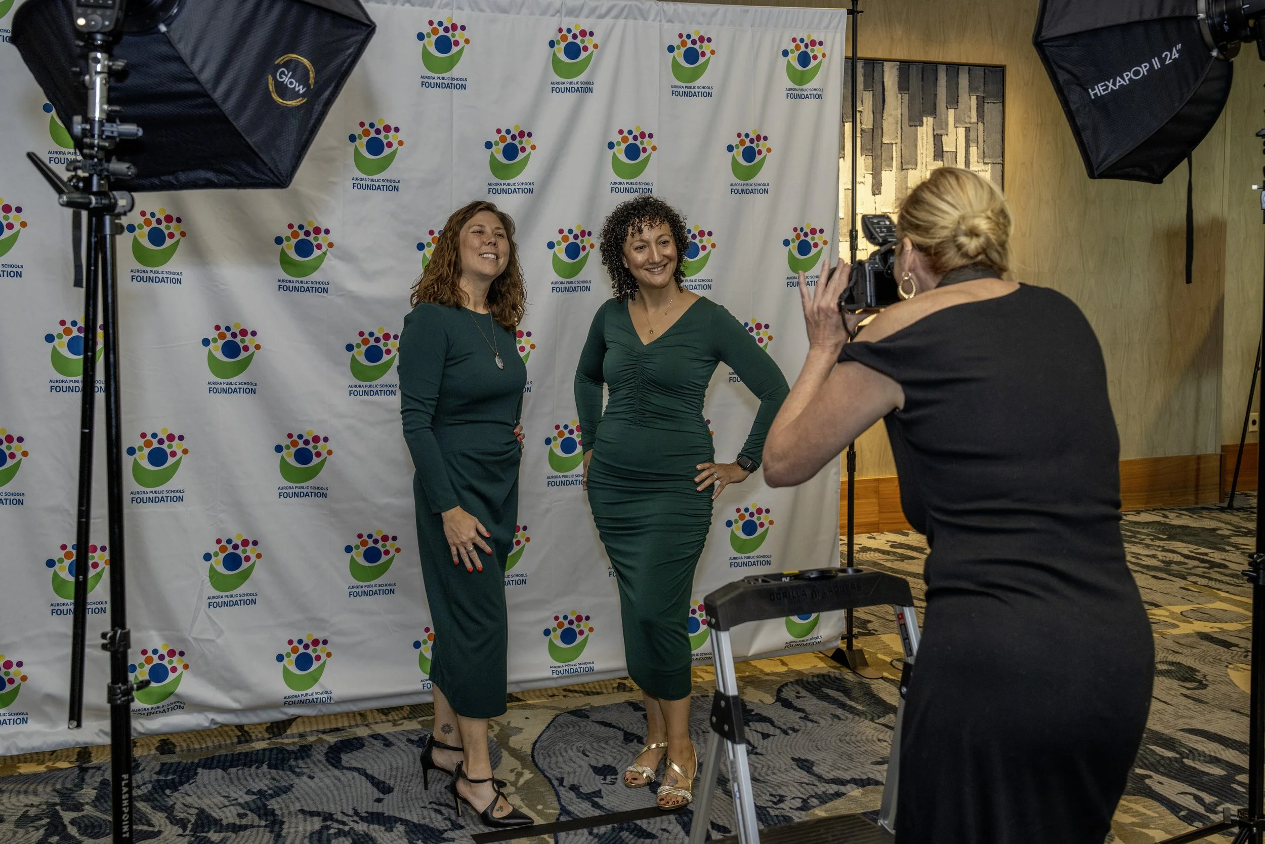 Two women in green dresses having their photo taken in front of a backdrop with the Aurora Public Schools Foundation logo, while a photographer captures the moment.
