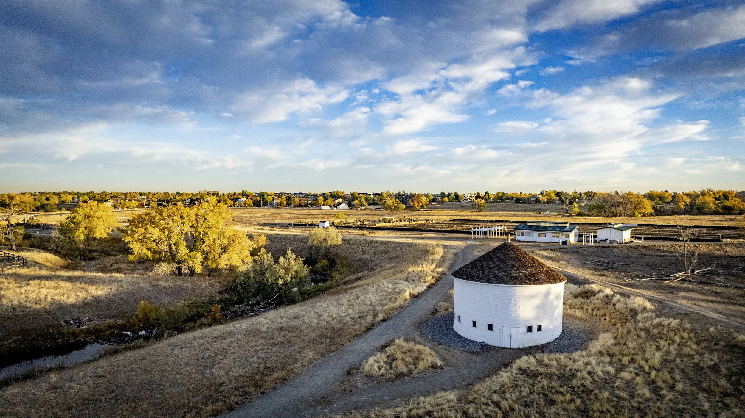 A rural landscape with a round white barn with a dark shingled roof, a gravel driveway, and dry grass. There are a few trees with yellow leaves and some farm buildings in the background under a partly cloudy sky.