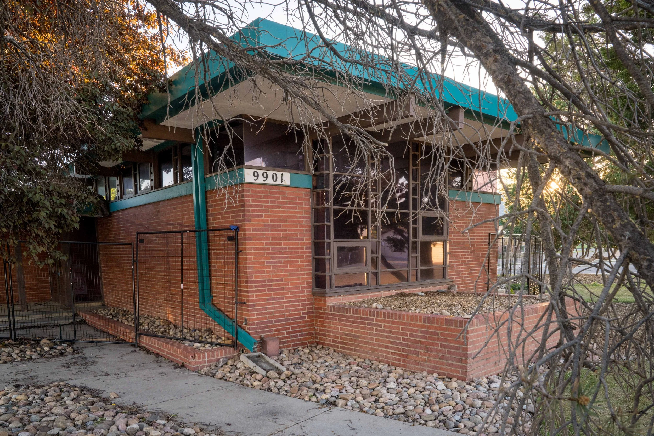 A brick house with a teal roof, large windows, and a brick planter box in front. The house number 9901 is visible, and there are leafless tree branches in the foreground.