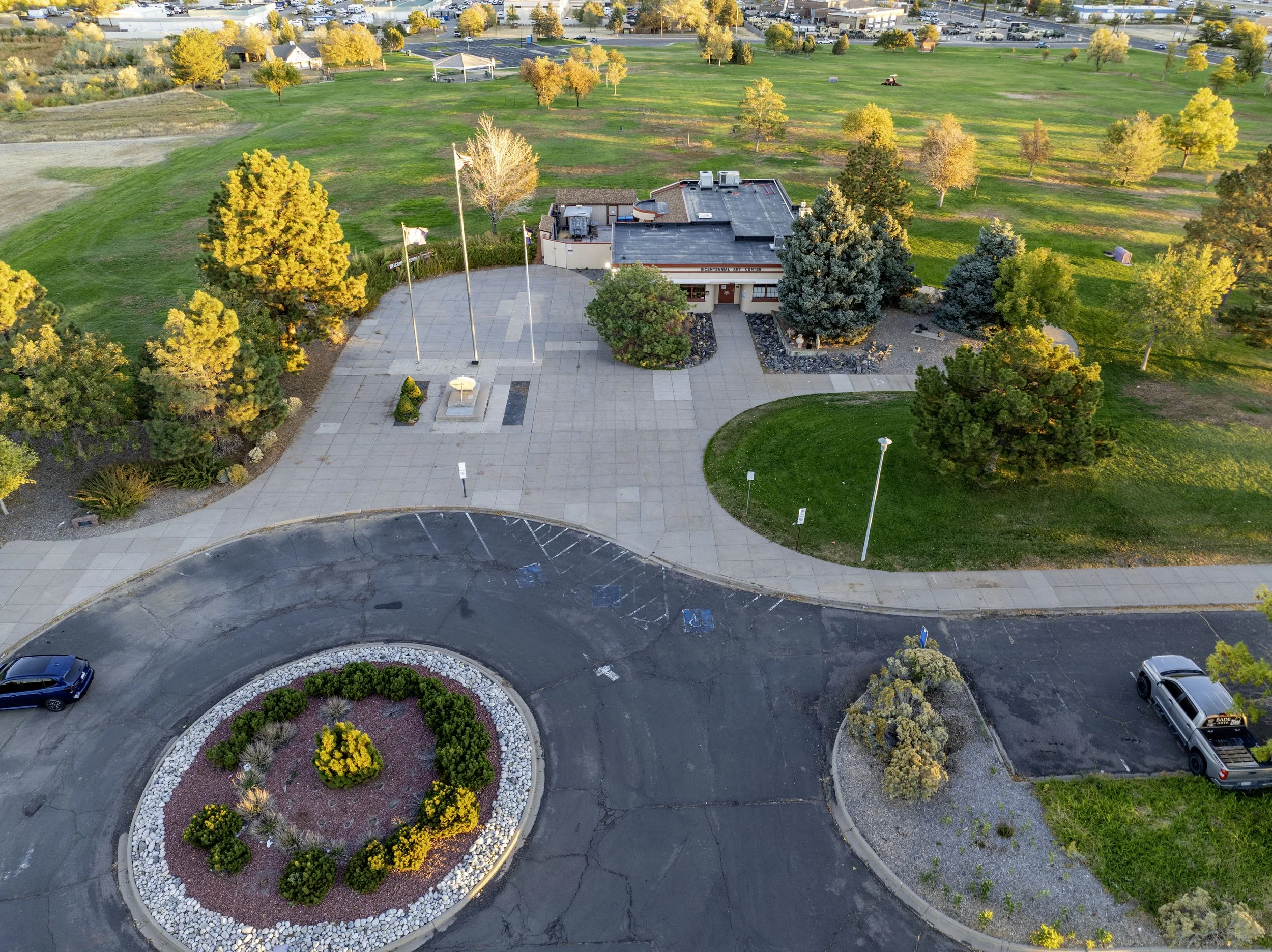An aerial view of a park with a parking lot, landscaped circular area with plants and yellow flowers, buildings, greenery, trees, and open fields during autumn.